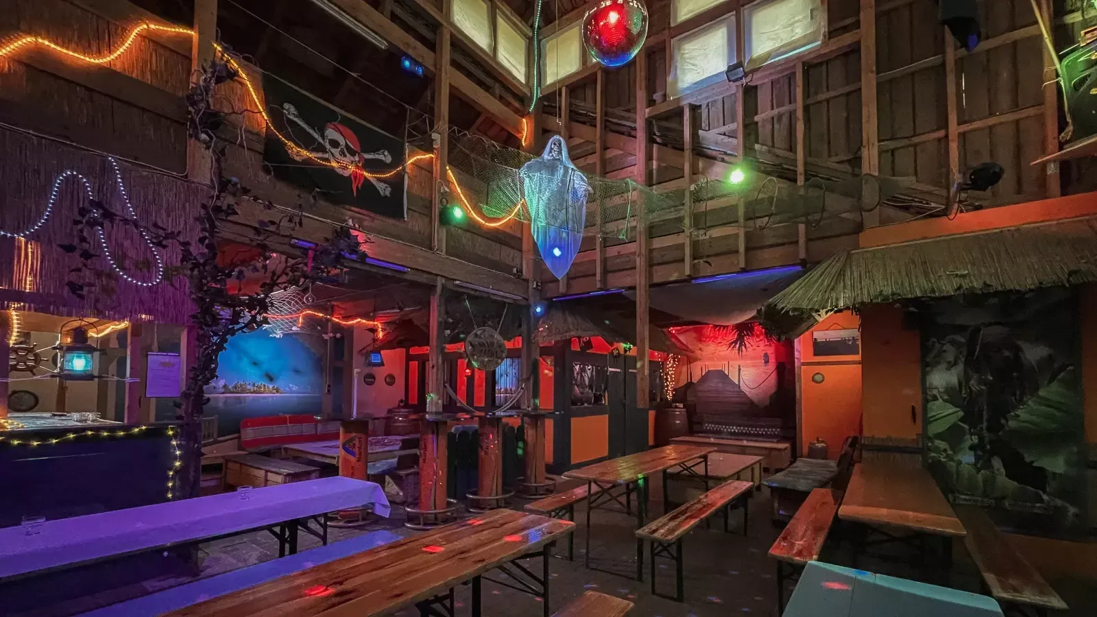 Interieur einer Themenbar mit Tiki-Dekor, Lichterketten, Piratenflagge und langen Tischen.
