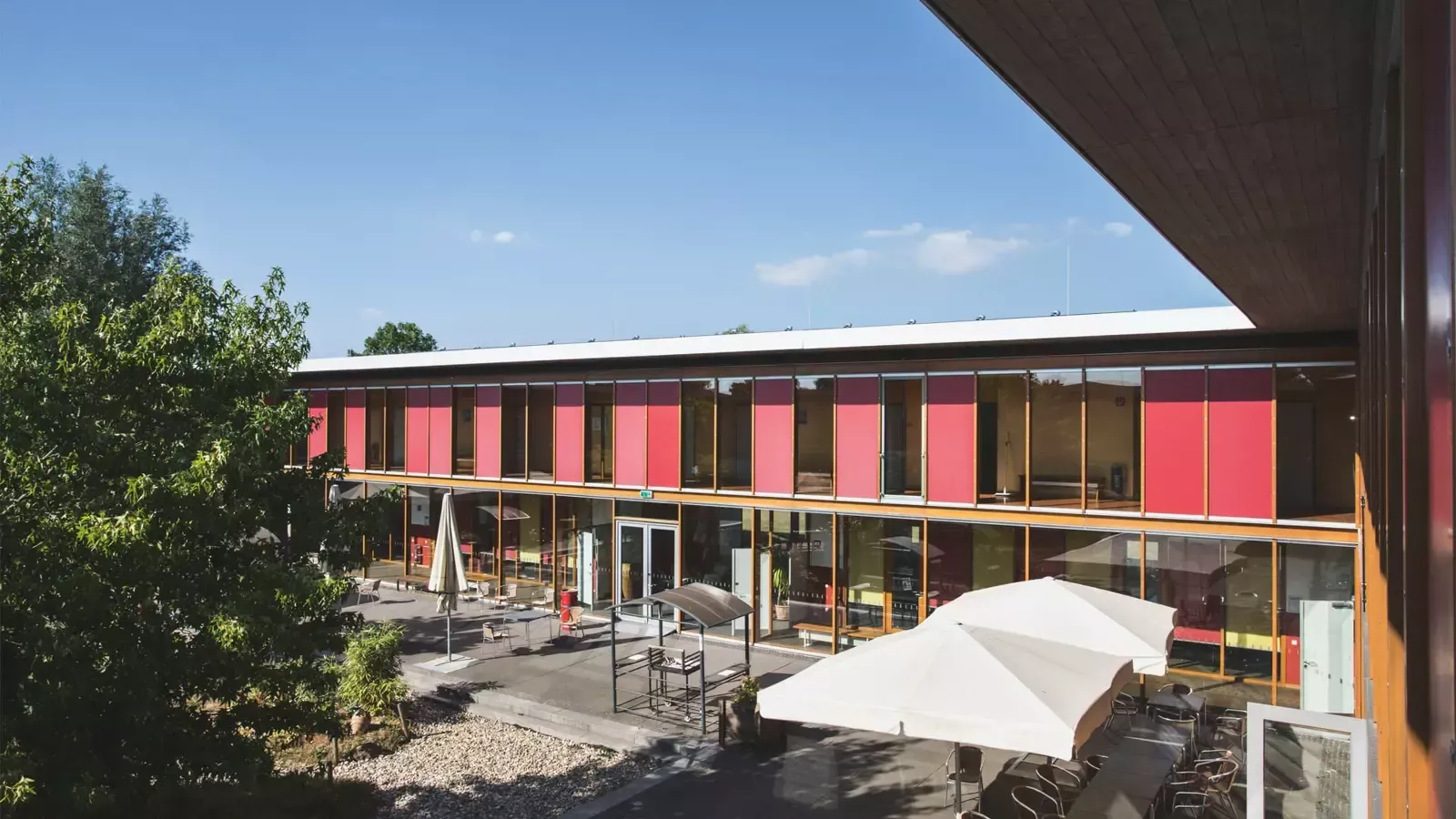 Außenaufnahme eines modernen Gebäudes mit roten Fensterfronten. Davor befindet sich eine Terrasse mit Sonnenschirmen und Tischen.