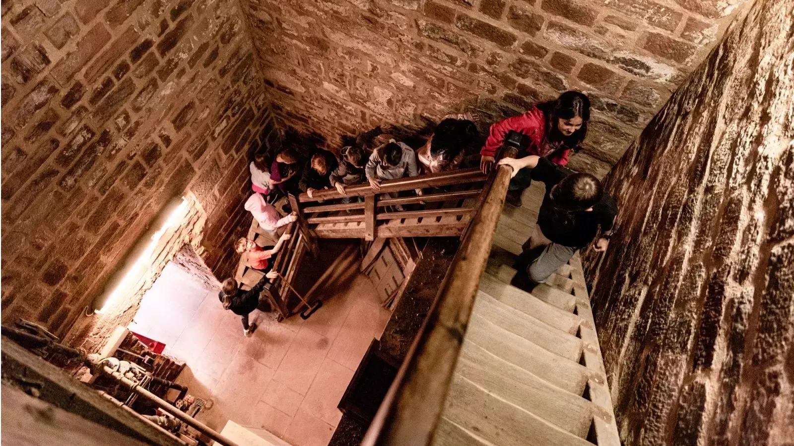 Menschen auf einer Holztreppe in einem Steinturm, die nach unten schauen.