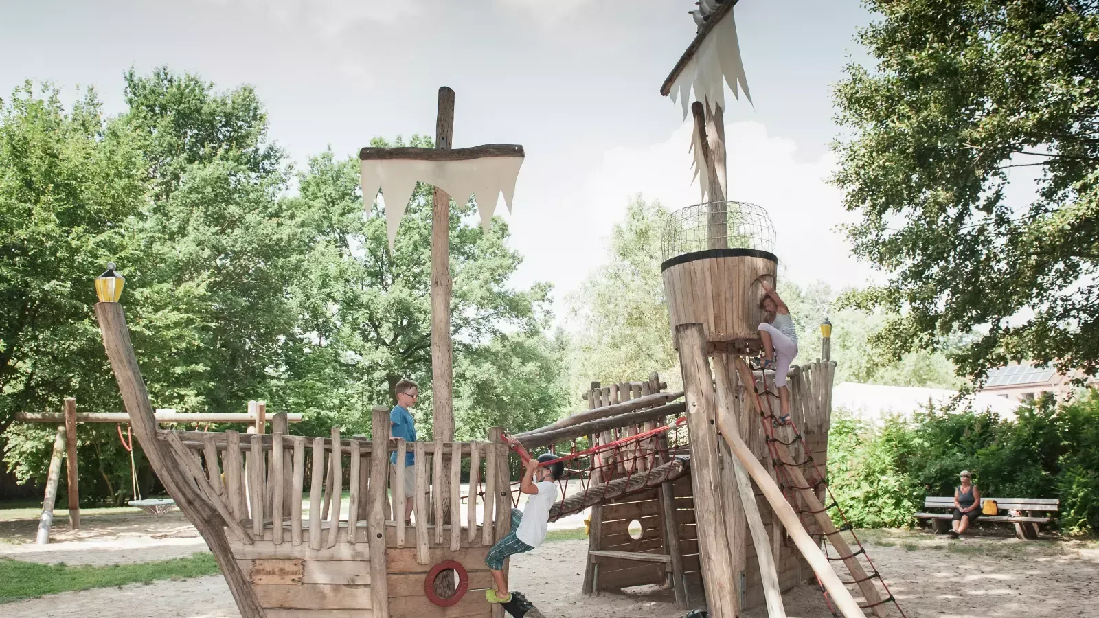 Spielplatz in Form eines Piratenschiffs aus Holz, auf dem Kinder klettern und spielen. Bäume im Hintergrund.