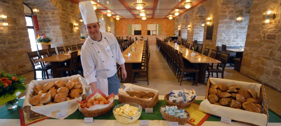 Ein Koch steht an einem Buffet mit Brotkörben und Speisen; im Hintergrund sind lange Esstische zu sehen.