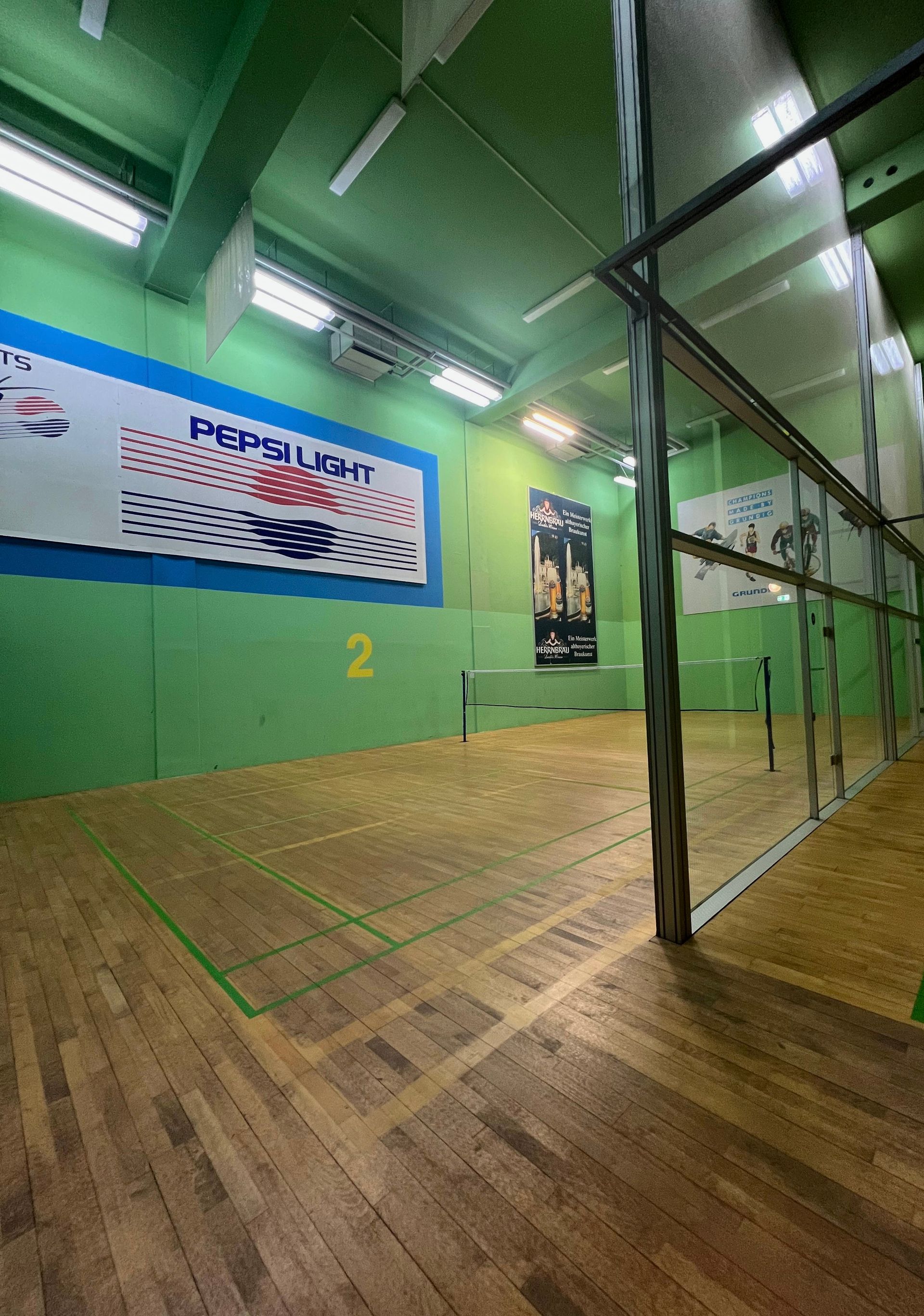 Squashcourt mit grünen Wänden, Holzboden und einem verglasten Zuschauerbereich.