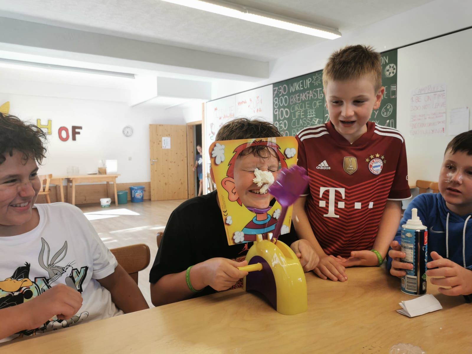 Vier Kinder spielen „Kuchengesichter“. Einem Kind ist Schlagsahne ins Gesicht geschmiert, die anderen schauen zu und lächeln. Die Szene spielt in einem Innenraum.