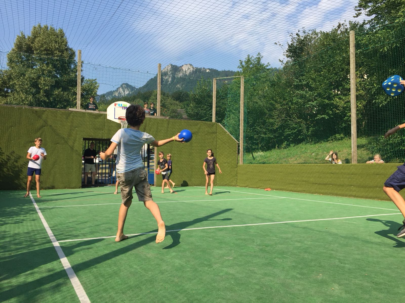 Kinder spielen Ball auf einem grünen Platz; im Hintergrund sind Berge zu sehen.