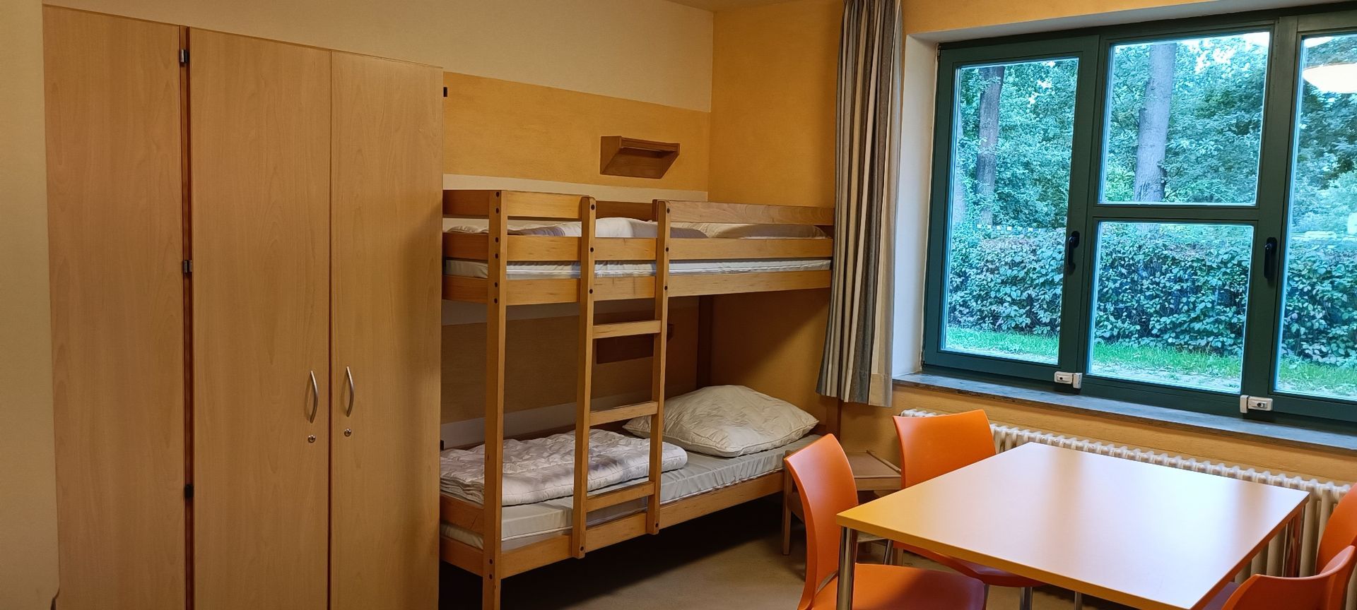 Ein Schlafsaal mit Etagenbetten, einem Tisch und einem großen Fenster mit Blick auf Bäume.