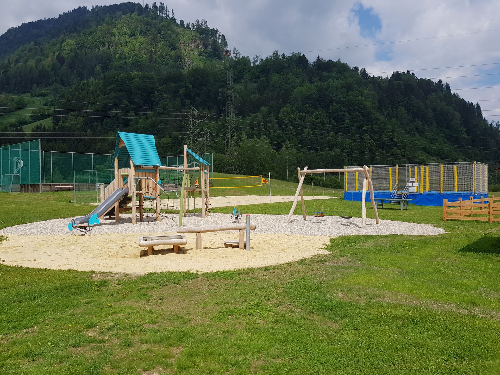 Spielplatz mit Schaukeln, Rutschen, Trampolin und Sandkasten vor einem Berg.