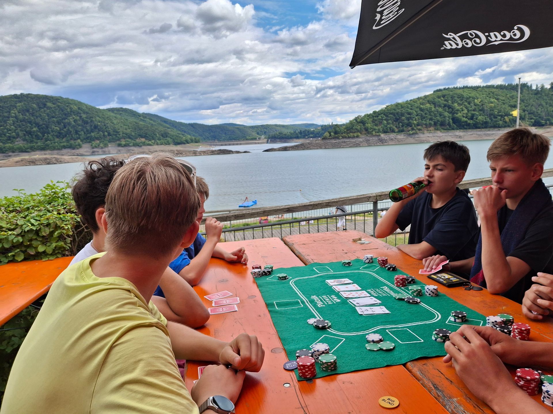 Eine Gruppe von Leuten spielt im Freien mit Blick auf den See Poker. Karten und Chips liegen auf dem Tisch.