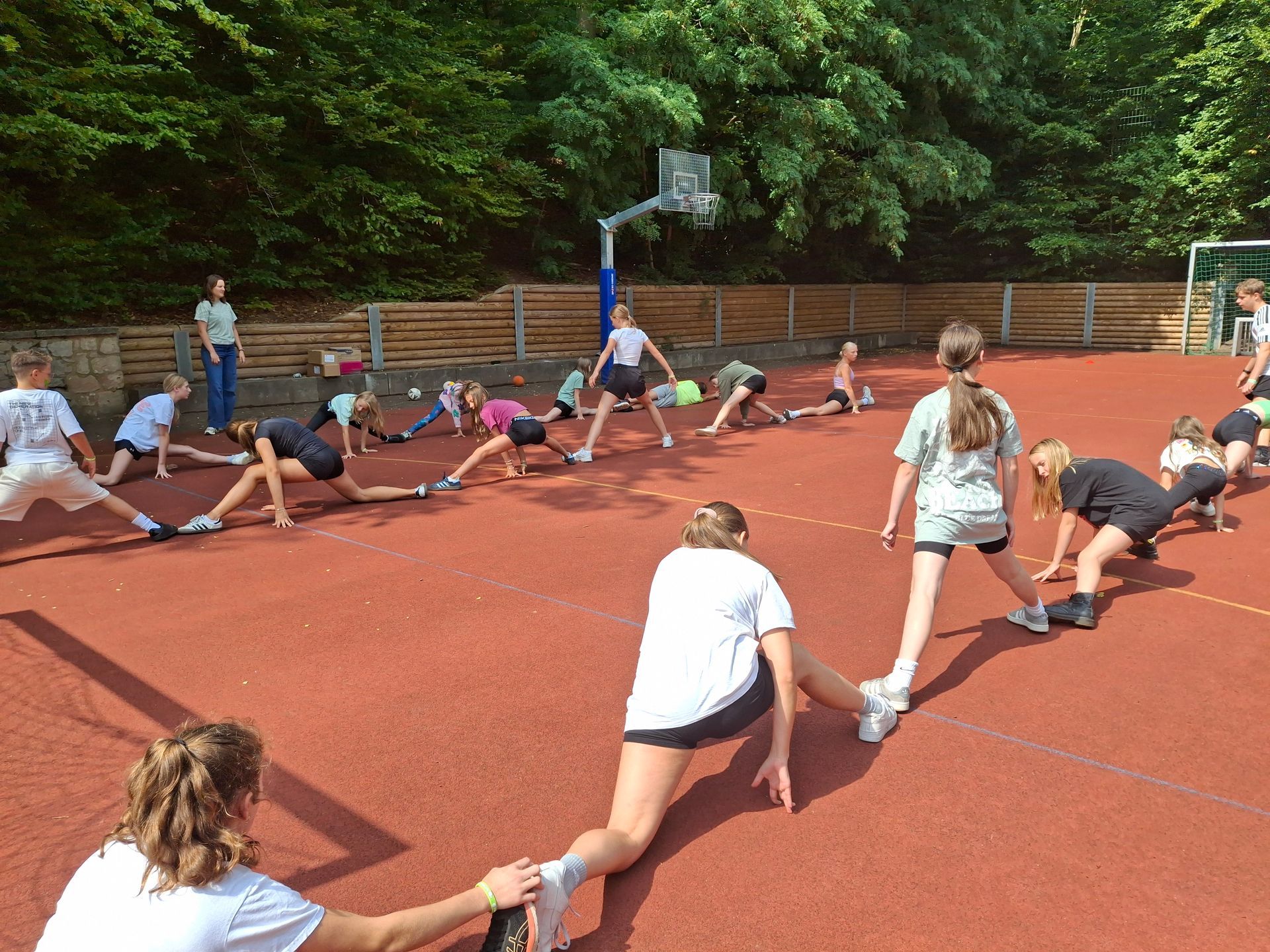 Eine Gruppe von Menschen dehnt sich auf einem roten Sportplatz unter einem Basketballkorb, umgeben von Bäumen.