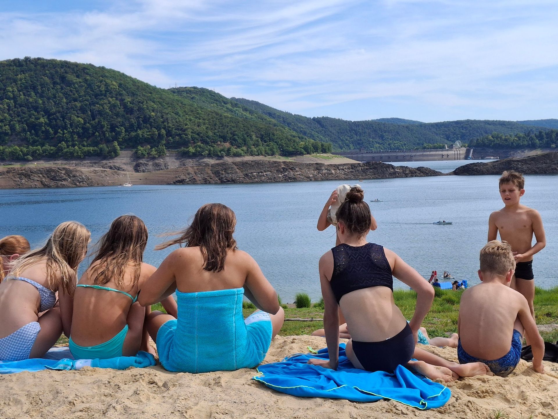 Menschen entspannen an einem Strand an einem See, im Hintergrund erheben sich Hügel. Sonniger Tag.