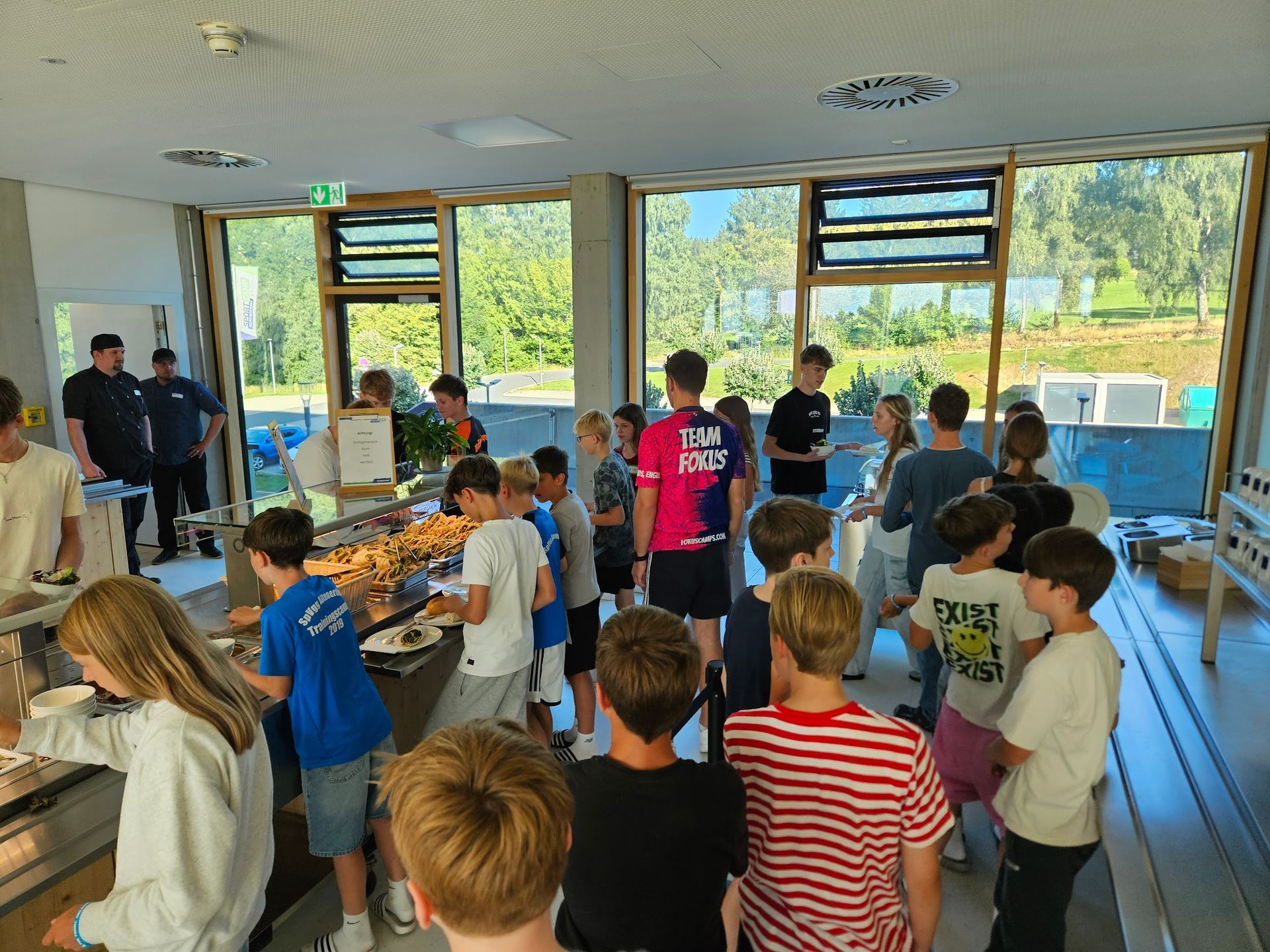 Kinder am Buffet, die sich Essen aussuchen. Andere stehen Schlange. Sonniger Raum mit Fenstern.