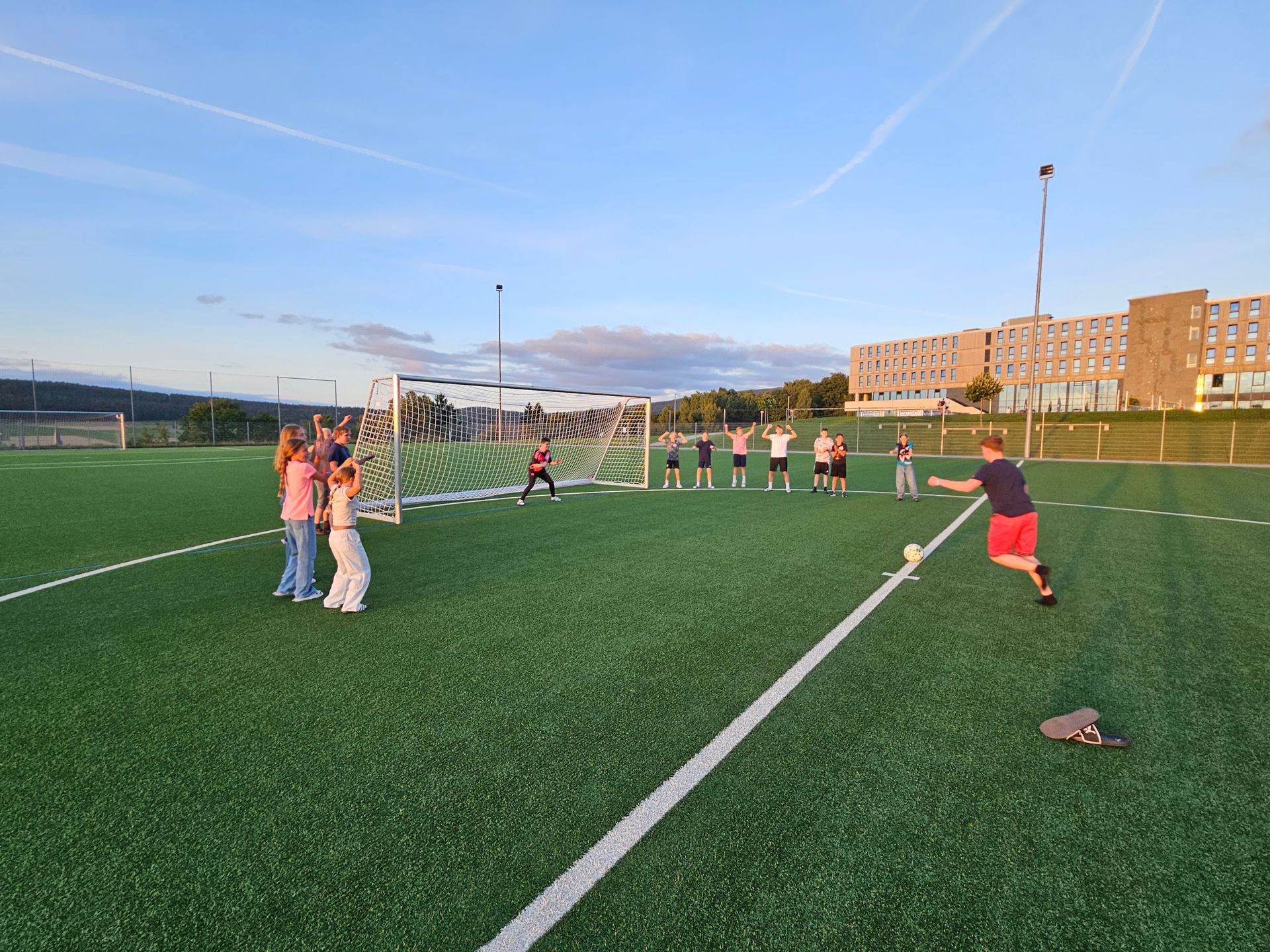 Menschen spielen Fußball auf einem grünen Rasen. Jemand schießt den Ball in Richtung Tor. Andere schauen zu.
