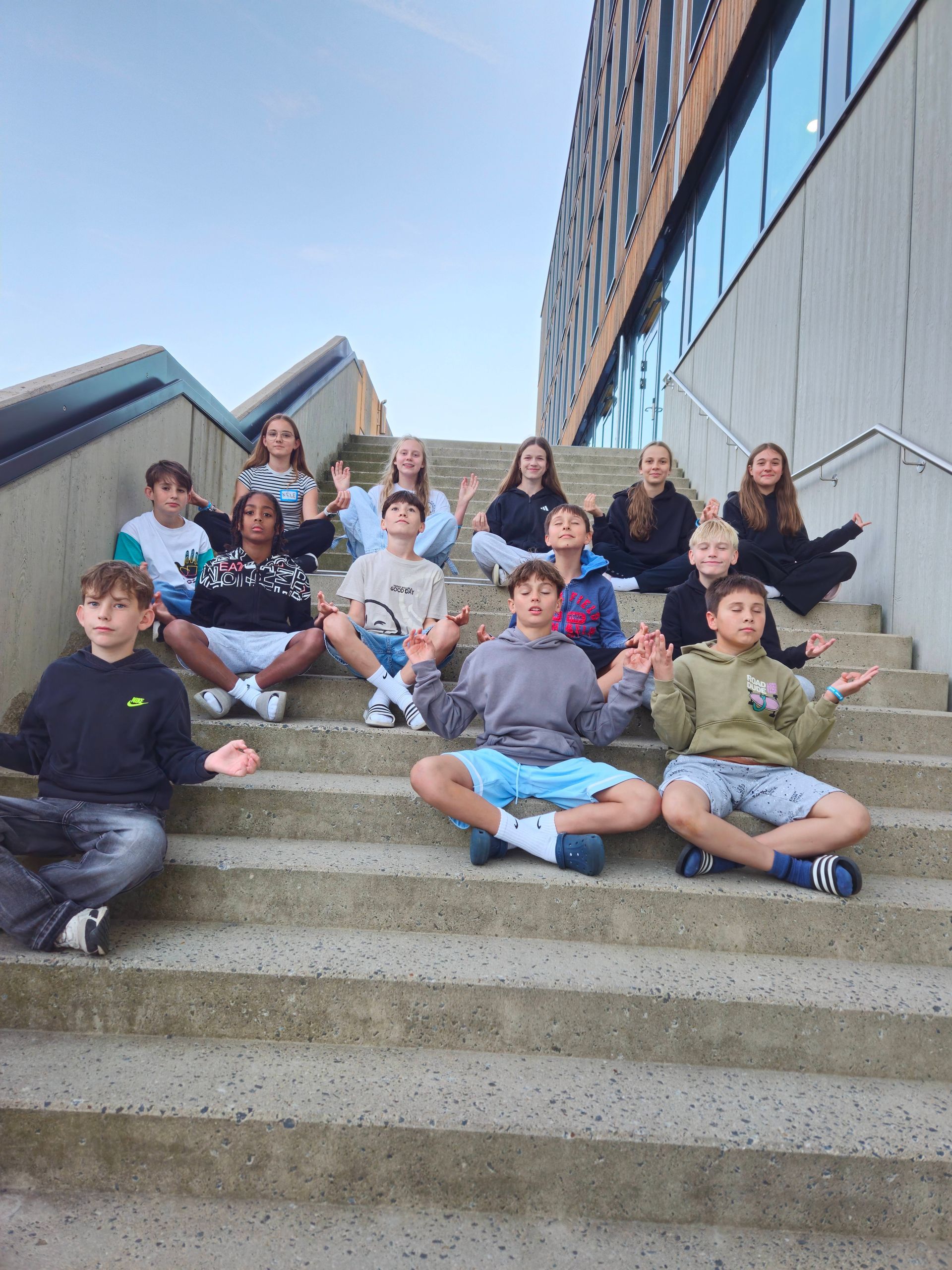 Eine Gruppe Kinder sitzt auf einer Betontreppe vor einem modernen Gebäude und hebt die Hände mit den Handflächen nach oben.