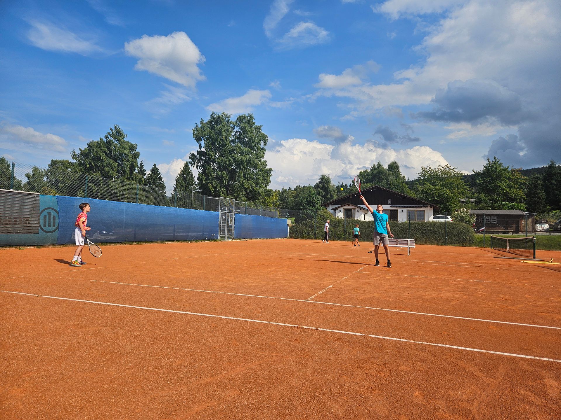 Zwei Personen spielen an einem sonnigen Tag Tennis auf einem Sandplatz.