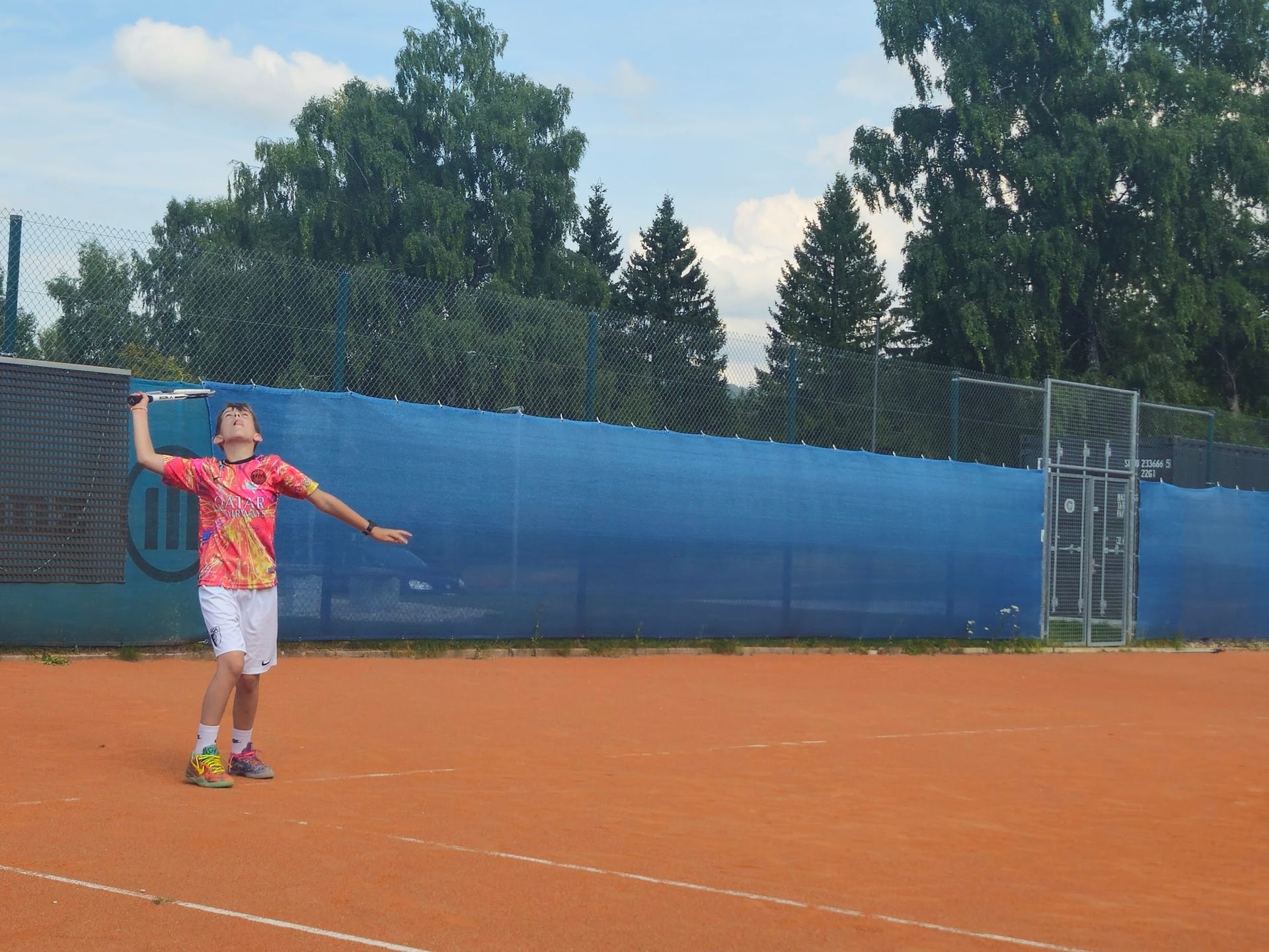 Ein Tennisspieler, der auf einem Sandplatz zum Aufschlag ansetzt, trägt ein farbenfrohes Hemd und weiße Shorts. Im Hintergrund ist ein blauer Zaun zu sehen.