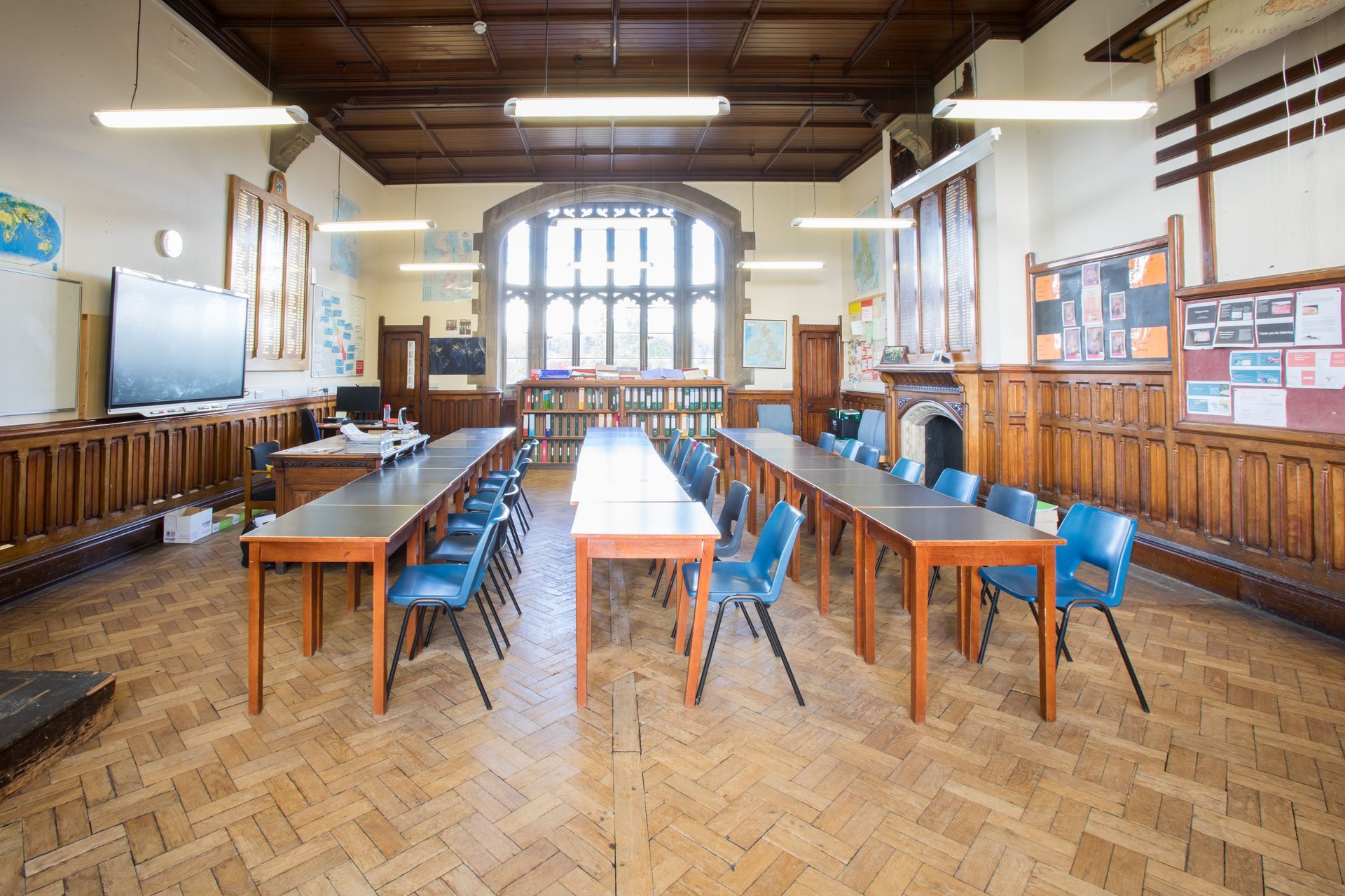 Leeres Klassenzimmer mit langen Schreibtischen, blauen Stühlen, kunstvoller Holzvertäfelung und großem Fenster.