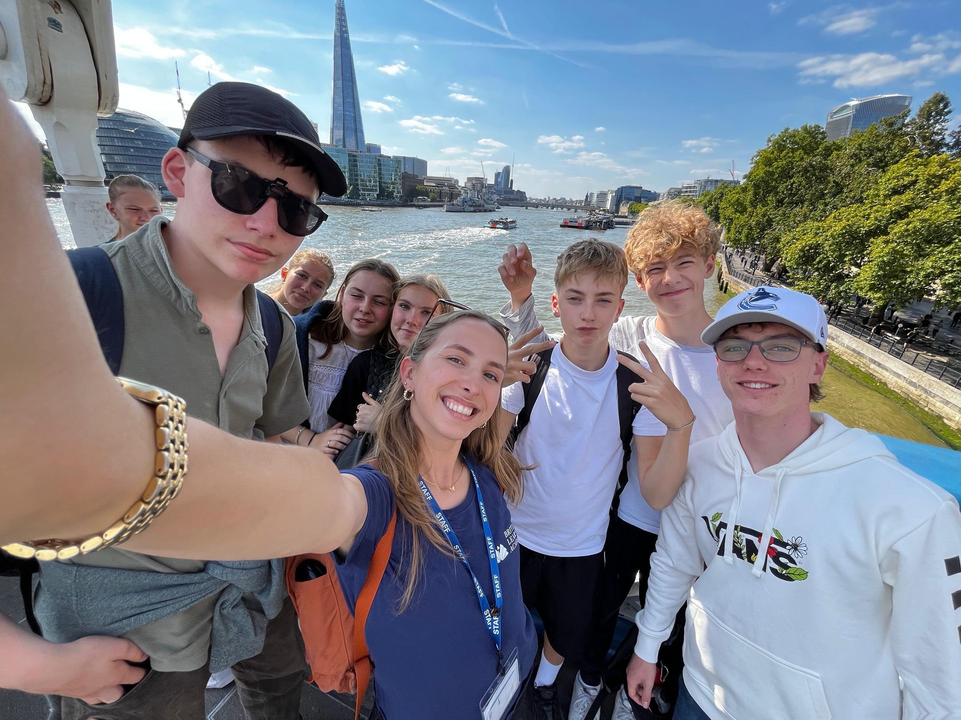 Eine Gruppe von Menschen macht ein Selfie auf einer Brücke, im Hintergrund ist The Shard zu sehen. Sonniger Tag.