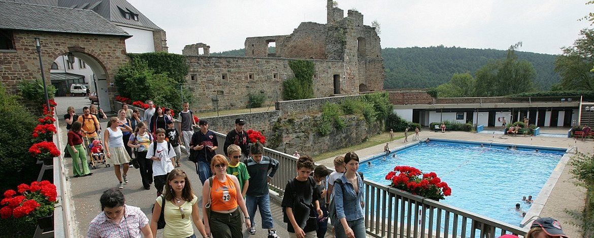 Menschen gehen in der Nähe eines Schwimmbads auf eine Burgruine zu.