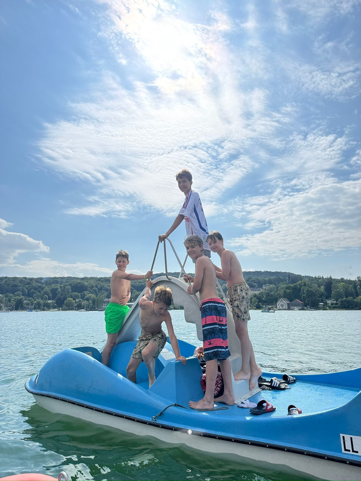 Eine Gruppe Jungen auf einem blauen Tretboot auf einem See unter bewölktem Himmel.