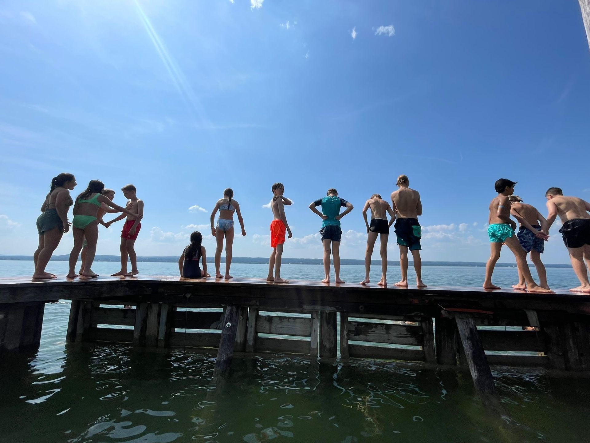 Menschen stehen auf einem Holzsteg und bereiten sich darauf vor, unter blauem Himmel in einen See zu springen.