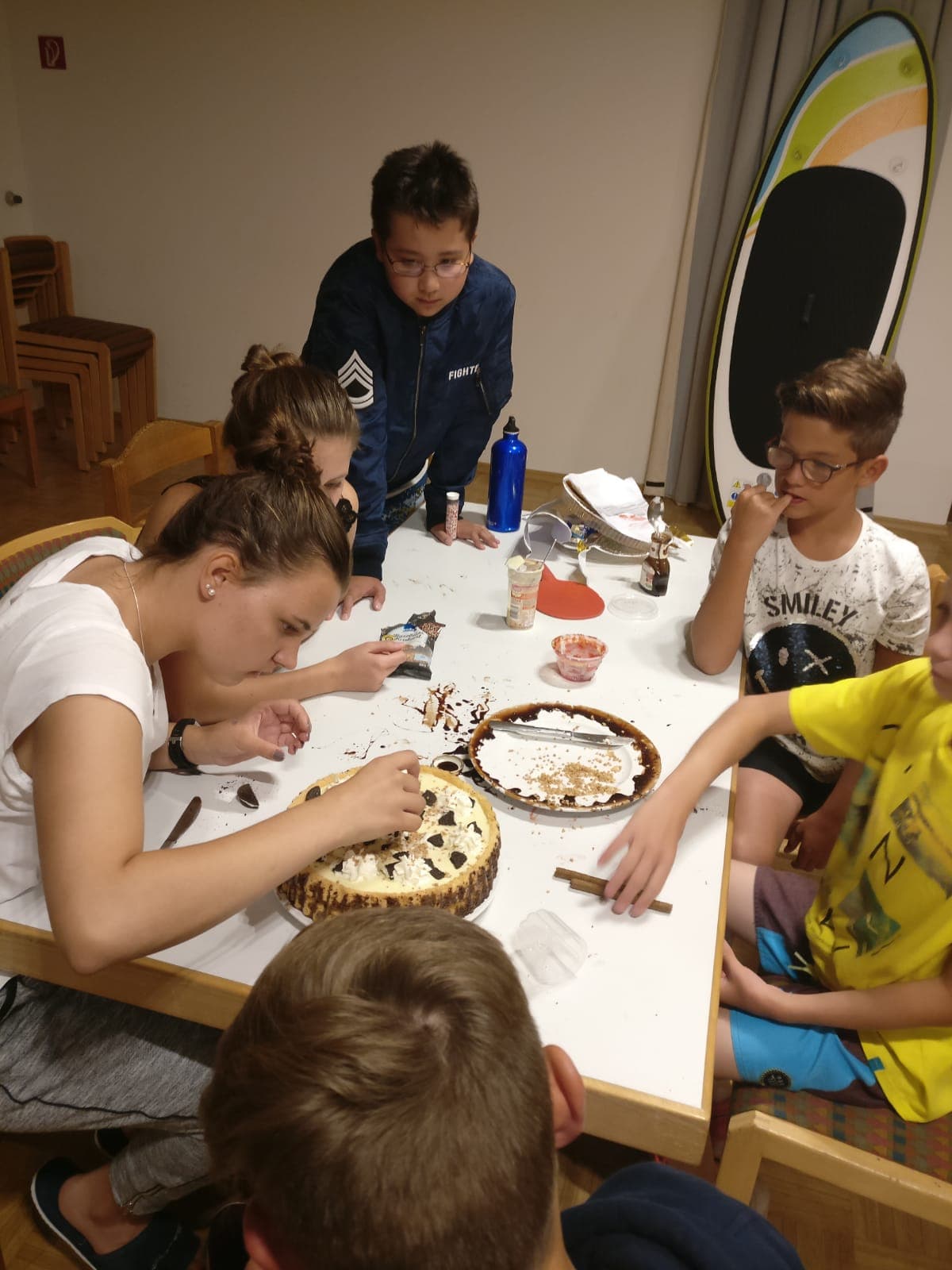 Eine Gruppe Kinder sitzt an einem Tisch, im Hintergrund ein Surfbrett