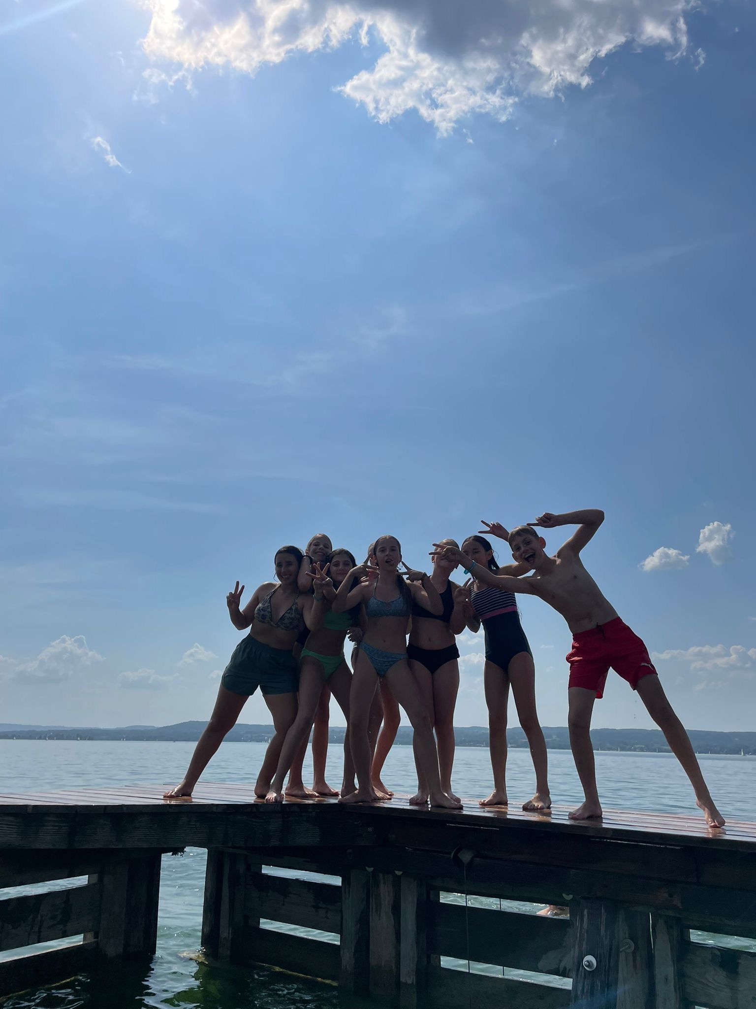 Eine Gruppe von Menschen in Badekleidung posiert auf einem Holzsteg über dem Wasser unter blauem Himmel.