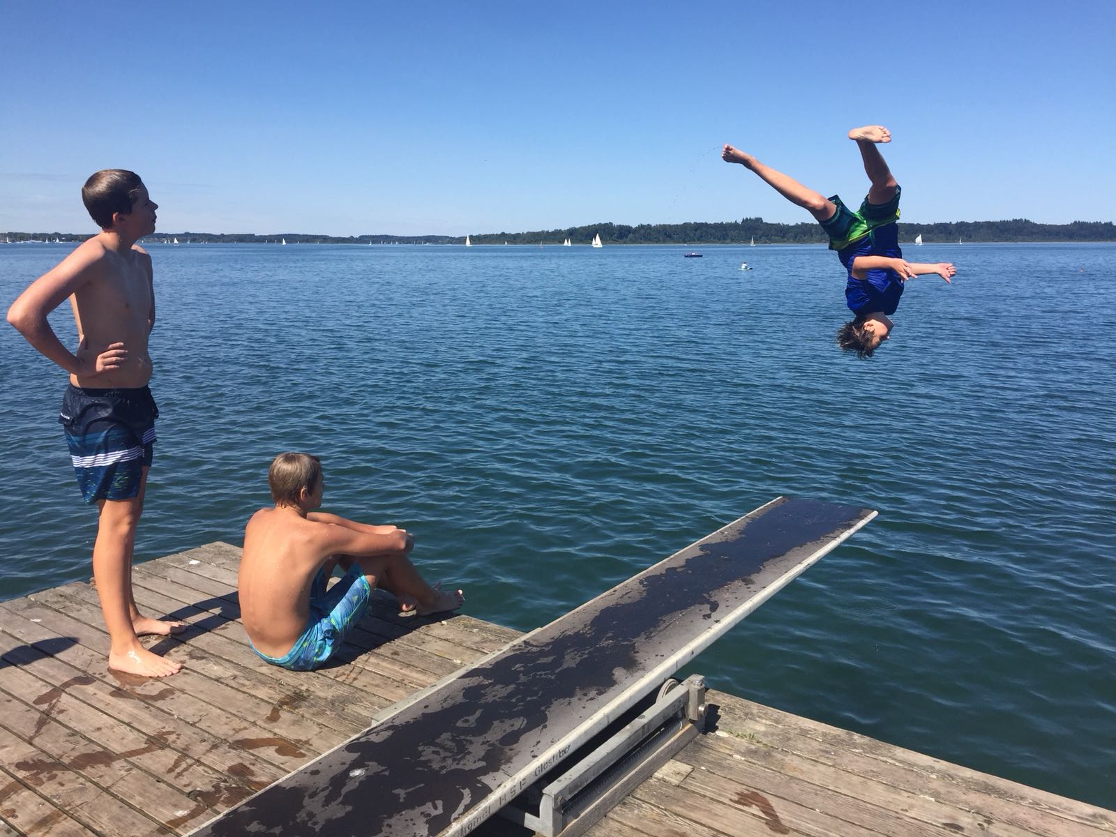 Drei Jungen an einem See: einer macht gerade einen Rückwärtssalto in der Luft, zwei andere schauen ihm von einem Steg aus zu. Sonniger Tag.