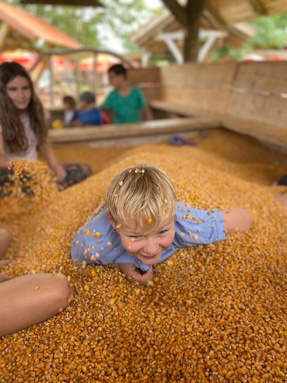 Ein Junge lächelt, während er in einem Außenspielbereich in Maiskörnern badet; andere Kinder sind zu sehen.