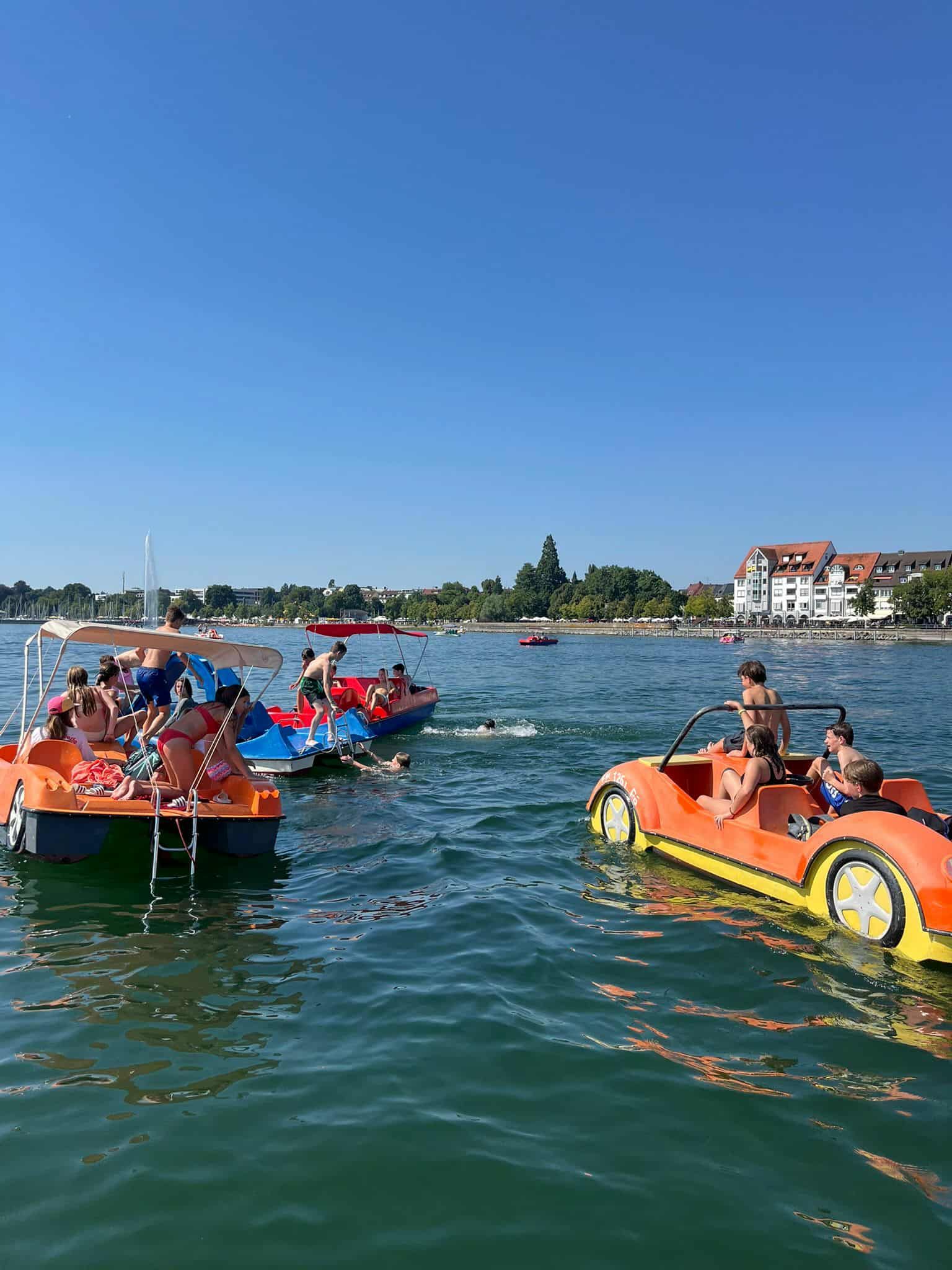 Leuchtend orange Tretboote auf einem See, auf dem Menschen den sonnigen Tag genießen. Gebäude und Bäume am Ufer.