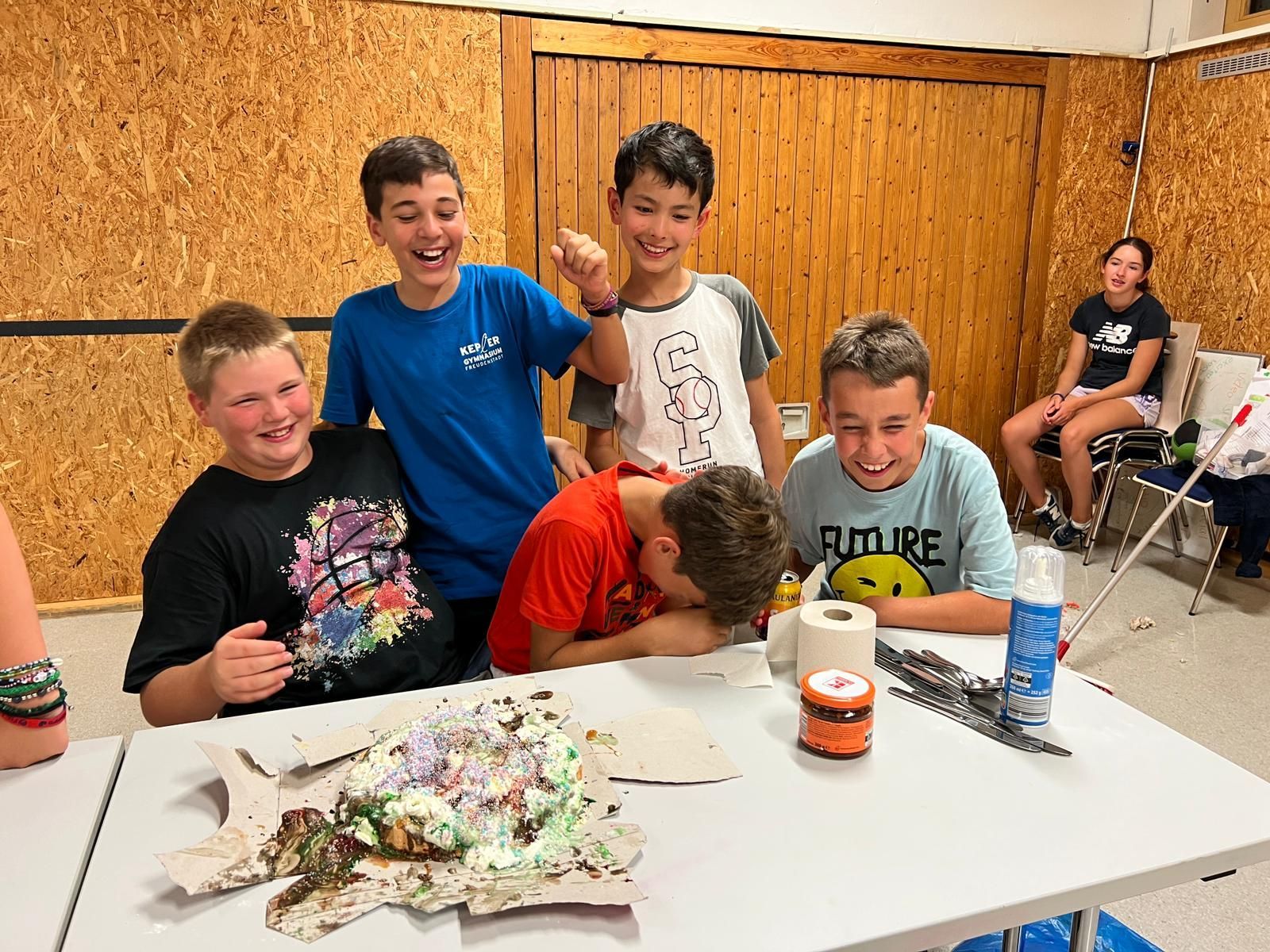 Fünf Kinder sitzen an einem Tisch, eines liegt mit dem Gesicht in einer Torte. Die anderen lachen und lächeln. Im Hintergrund eine Holzwand.