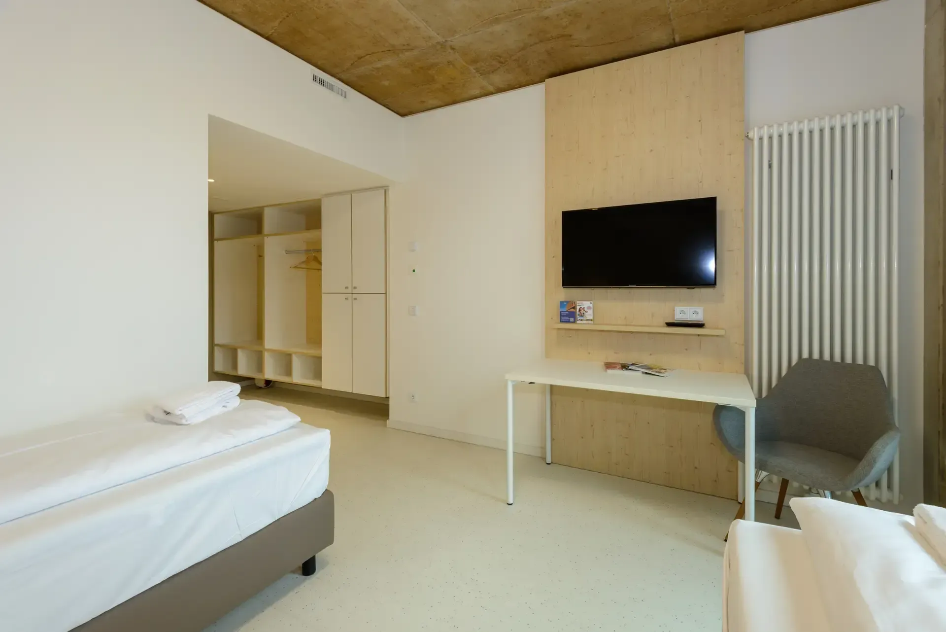 Hotelzimmer mit zwei Betten, Schreibtisch, Fernseher, Kleiderschrank und Heizkörper. Helle, moderne Einrichtung.