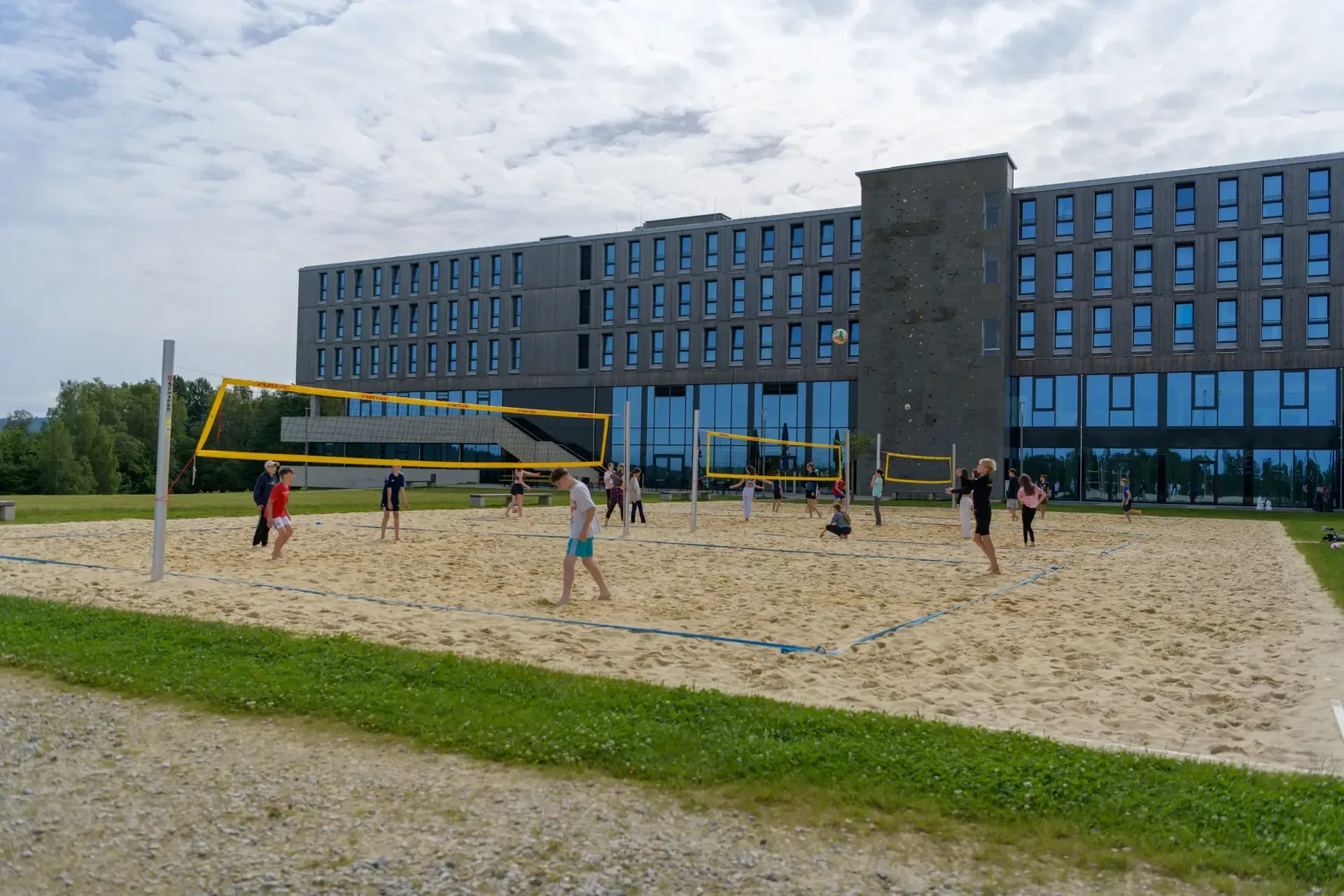 Menschen spielen Volleyball auf einem Sandplatz vor einem modernen Gebäude.