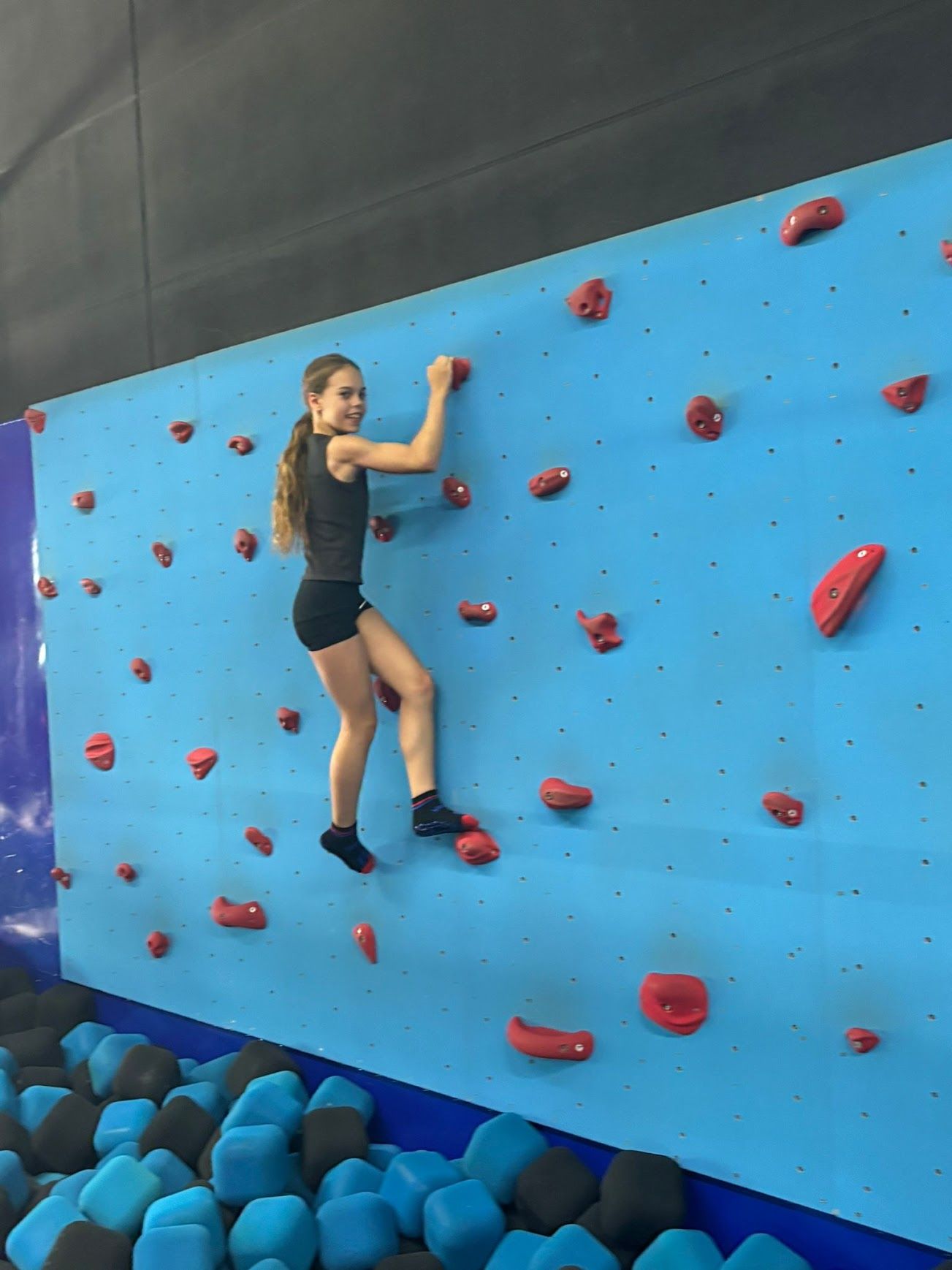 Ein junger Mensch klettert in einer Kletterhalle an einer blauen Felswand mit roten Griffen.