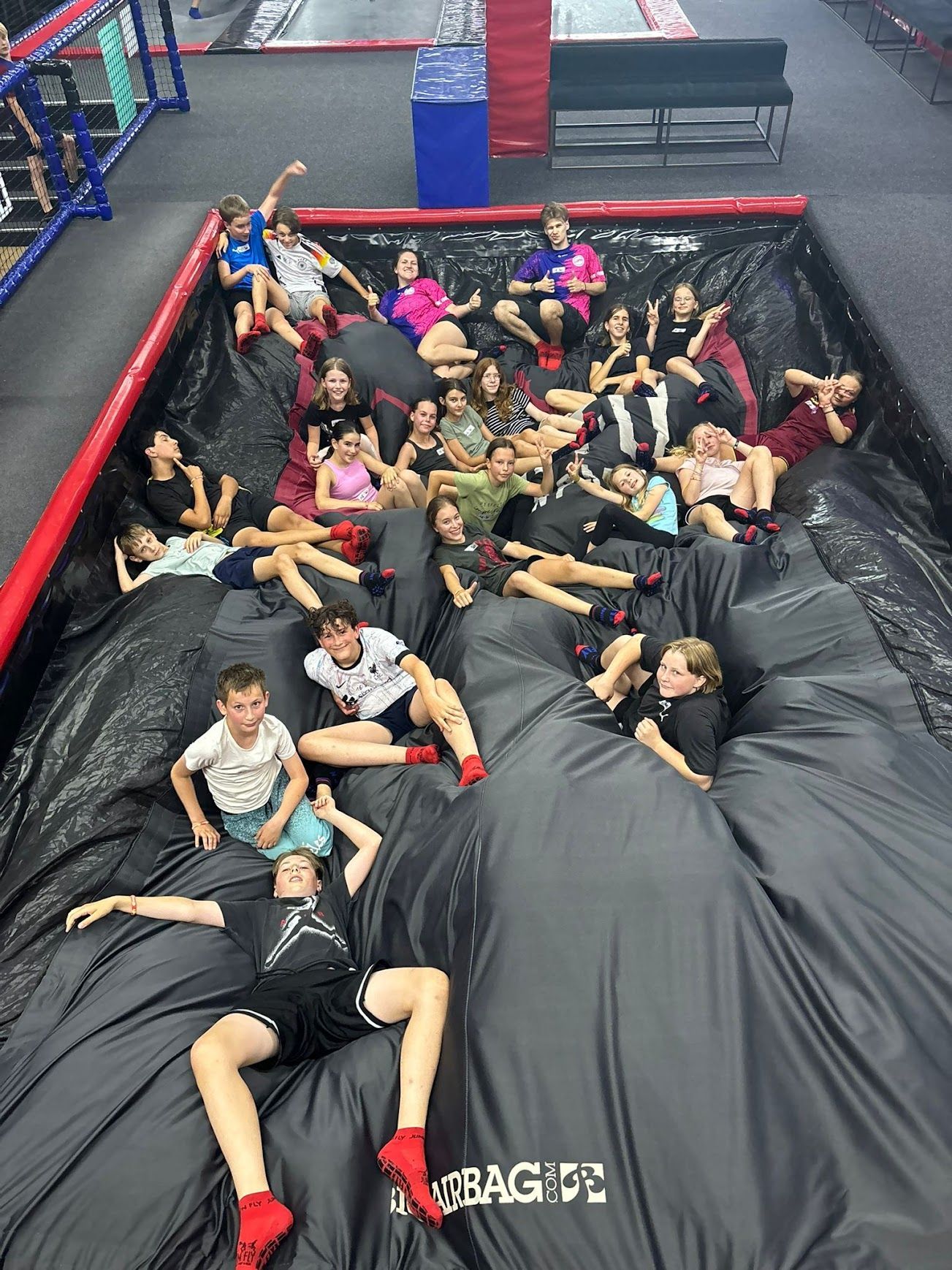 Eine Gruppe von Menschen sitzt auf einem großen schwarzen Luftkissen. Einige lächeln. Die Szene spielt in einem Trampolinpark.