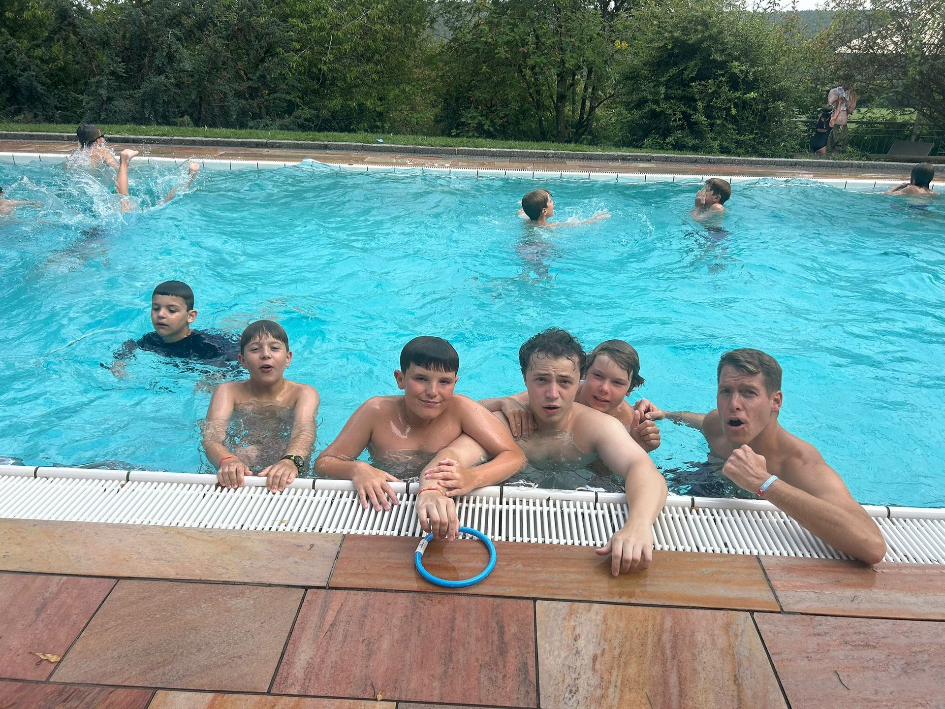 Eine Gruppe Kinder posiert mit aufgeregten Gesichtsausdrücken am Beckenrand eines Schwimmbeckens.