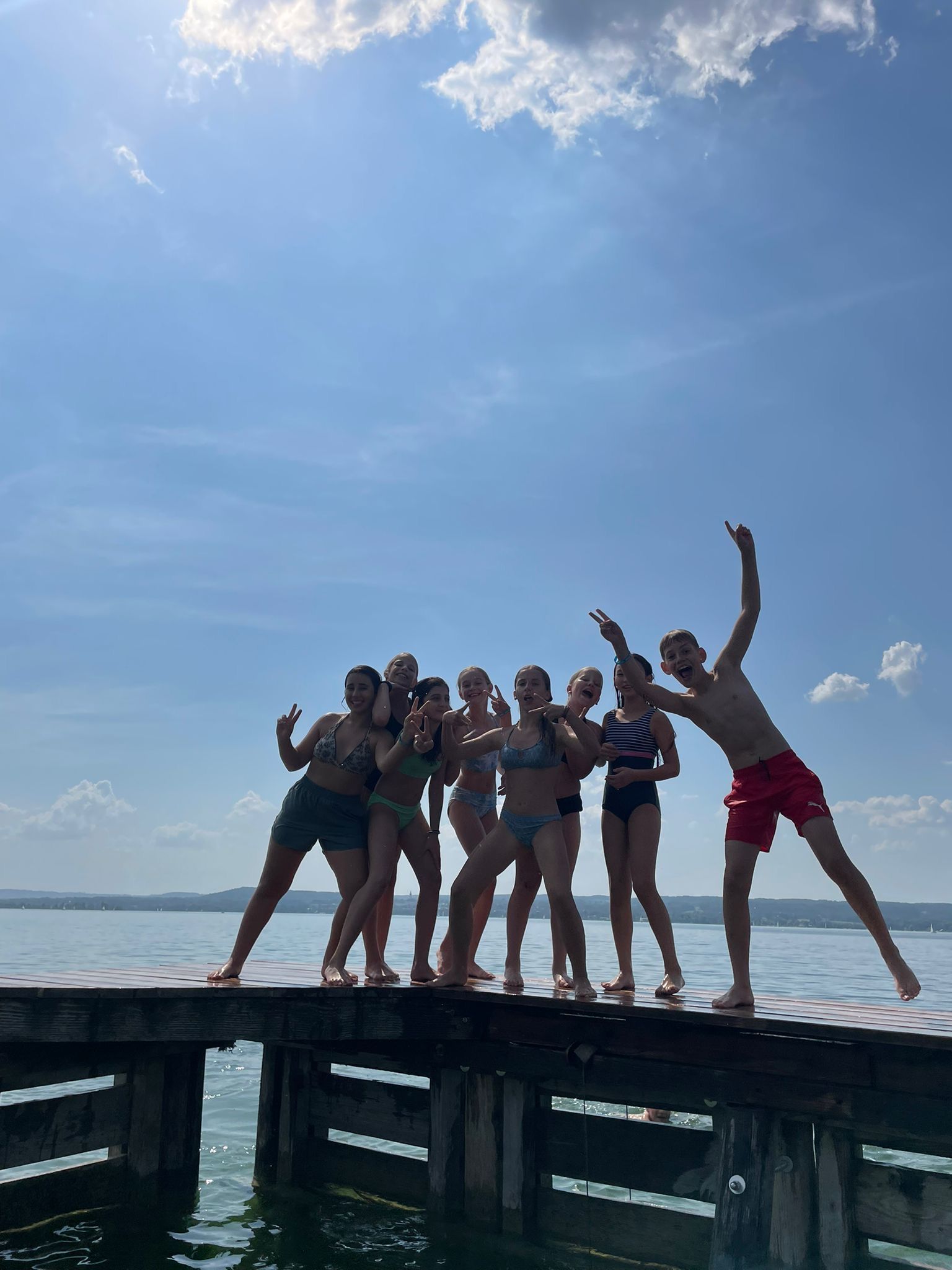 Eine Gruppe von Menschen in Badekleidung posiert an einem sonnigen Tag auf einem Holzsteg an einem See.