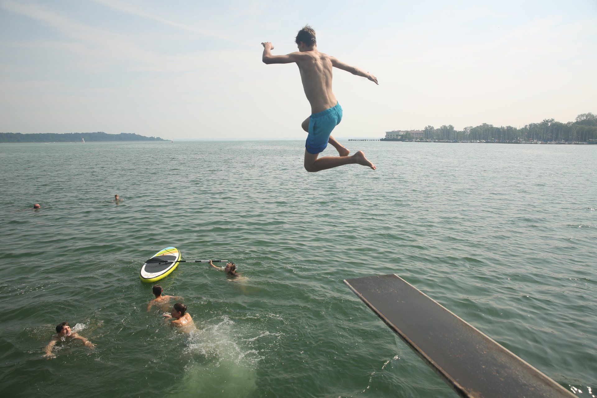 Eine Person springt von einem Sprungbrett in einen See. Andere schwimmen im Wasser; bewölkter Tag.