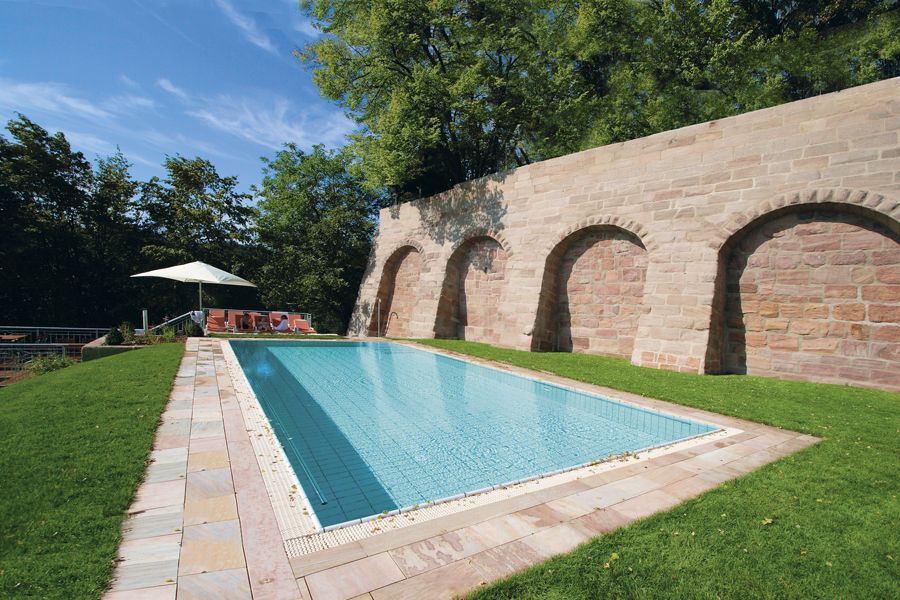 Rechteckiger Pool mit blauem Wasser neben einer Backsteinmauer mit Bogenöffnungen, umgeben von Gras und sonnigem Himmel.