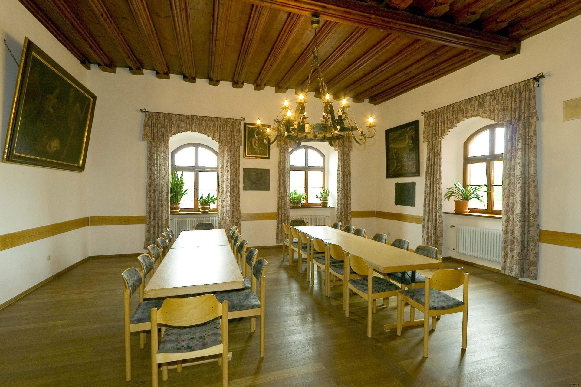 Ein Esszimmer mit langen Tischen, Stühlen und einem Kronleuchter. Fenster mit Vorhängen und Kunstwerken. Holzdecke und Holzböden.