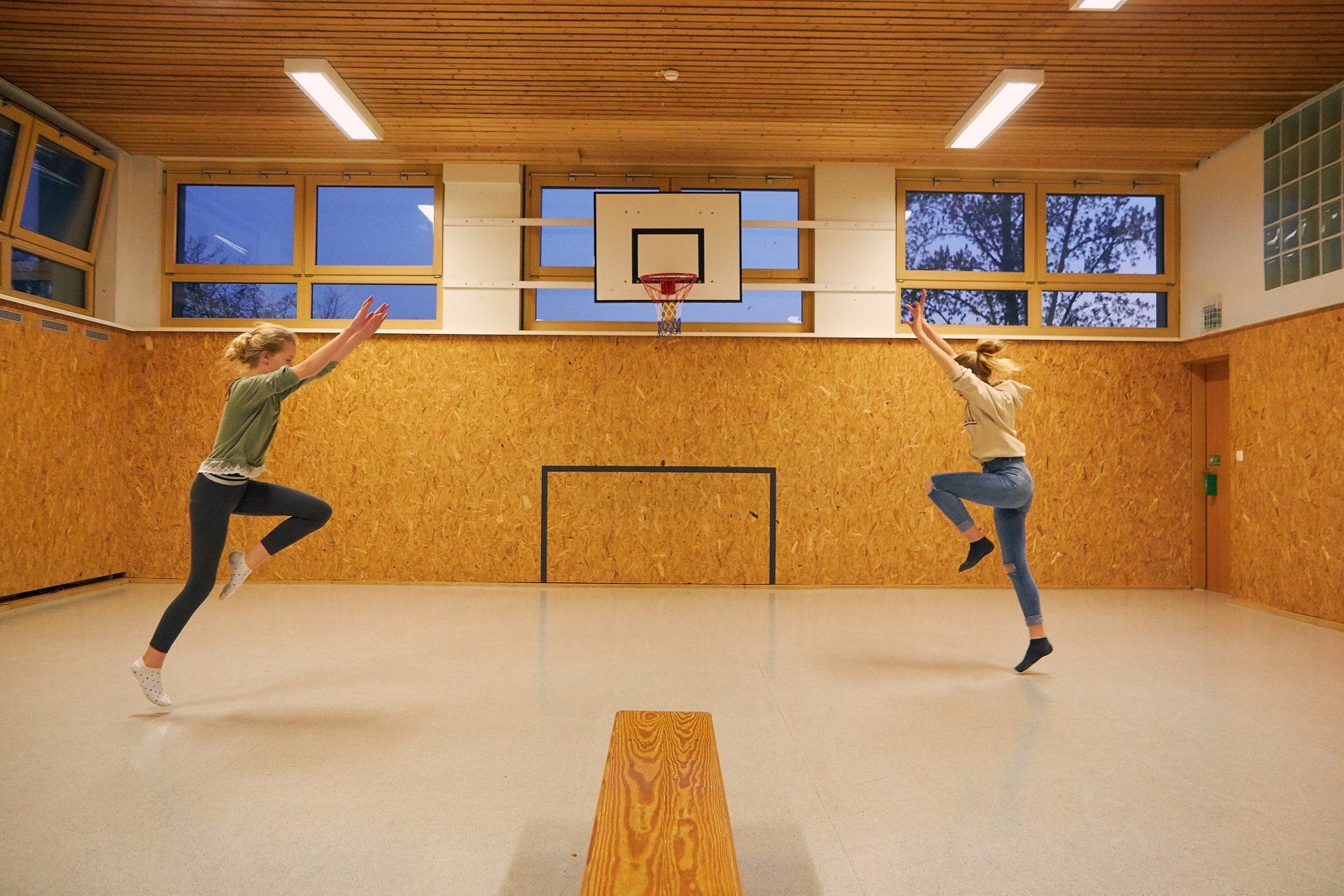 Zwei Personen springen in einer holzgetäfelten Turnhalle auf einen Basketballkorb zu.