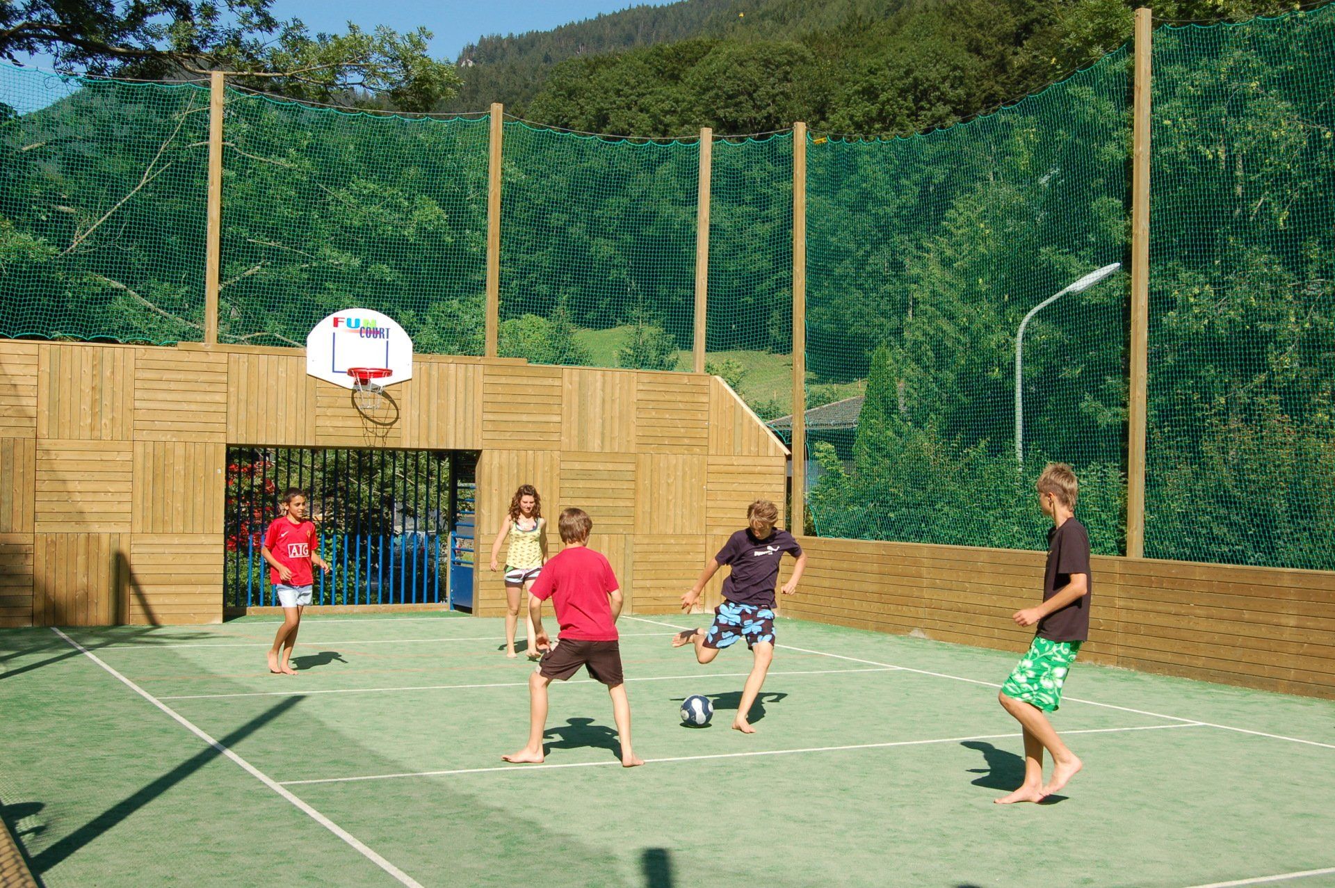 Kinder spielen Fußball und Basketball auf einem grünen Freiplatz mit Holzwänden und Netz.