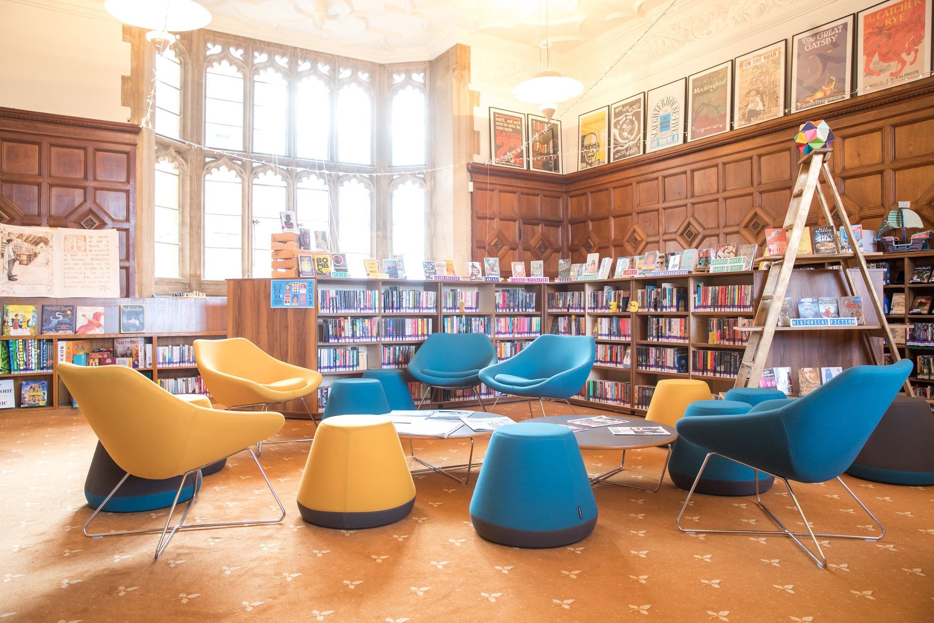 Gemütliches Bibliotheksinterieur mit Bücherregalen, Sitzecke (gelbe, blaue Stühle/Hocker) und großen Fenstern.