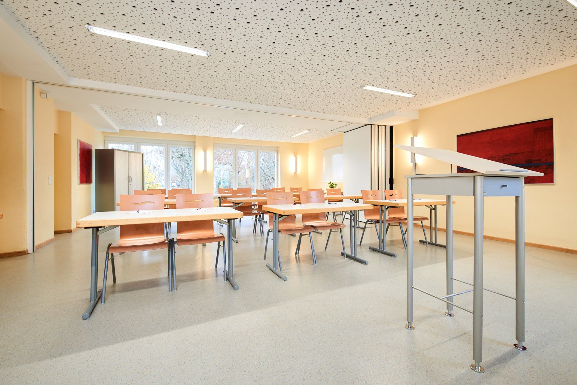 Klassenzimmer mit Reihen von Schulbänken, Rednerpult und Fenstern. Beige Wände, heller Boden, rote Bilder.