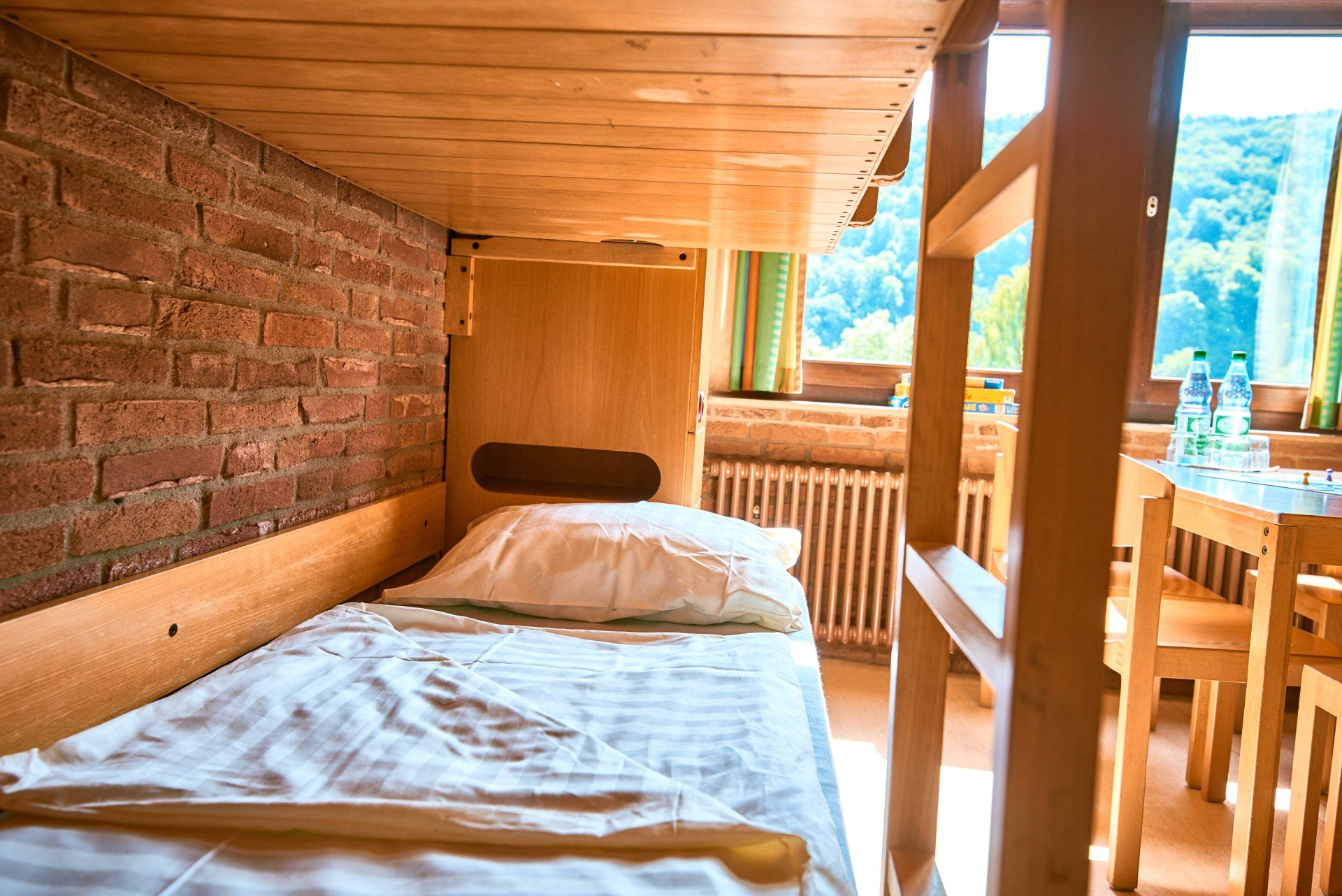 Etagenbett in einem rustikalen Zimmer mit freiliegender Ziegelwand, Fenster mit Blick auf einen Wald und Holzmöbeln.
