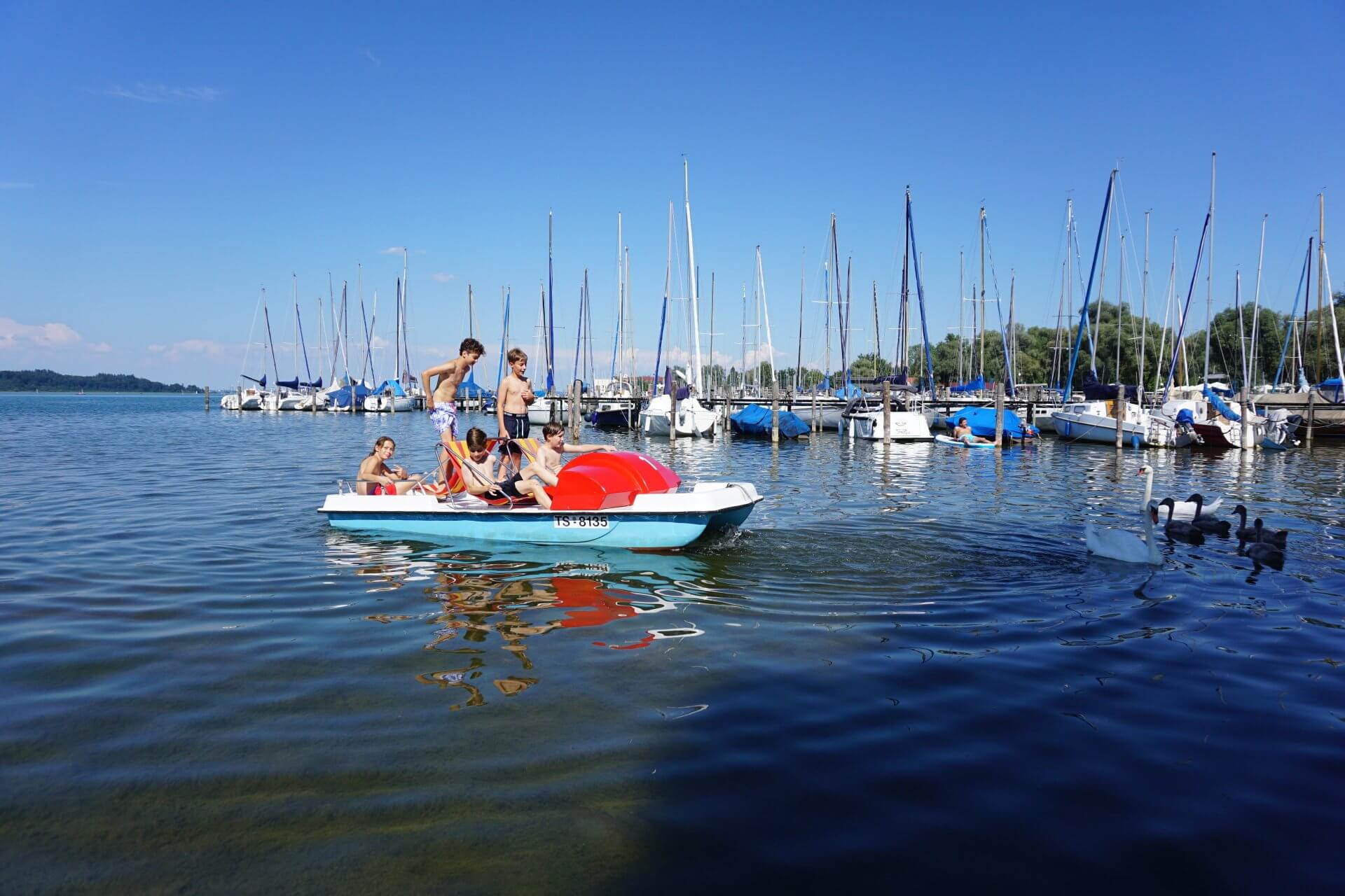 Menschen auf einem Tretboot, blaues Wasser, vor Anker liegende Segelboote, sonniger Tag.