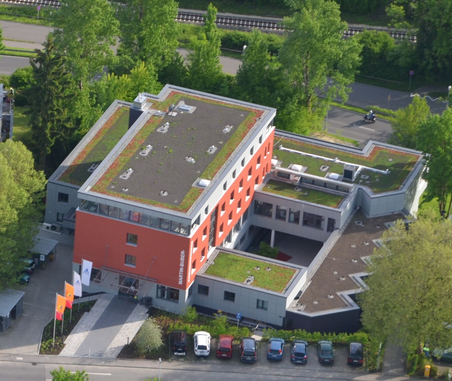 Luftaufnahme eines modernen Hotelgebäudes mit roter Fassade, grünem Dach und Parkplatz.
