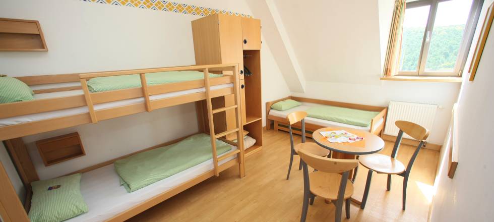 Ein Schlafsaal mit Etagenbetten, Einzelbett, kleinem Tisch und Stühlen. Holzmöbel und hellgrüne Bettwäsche.