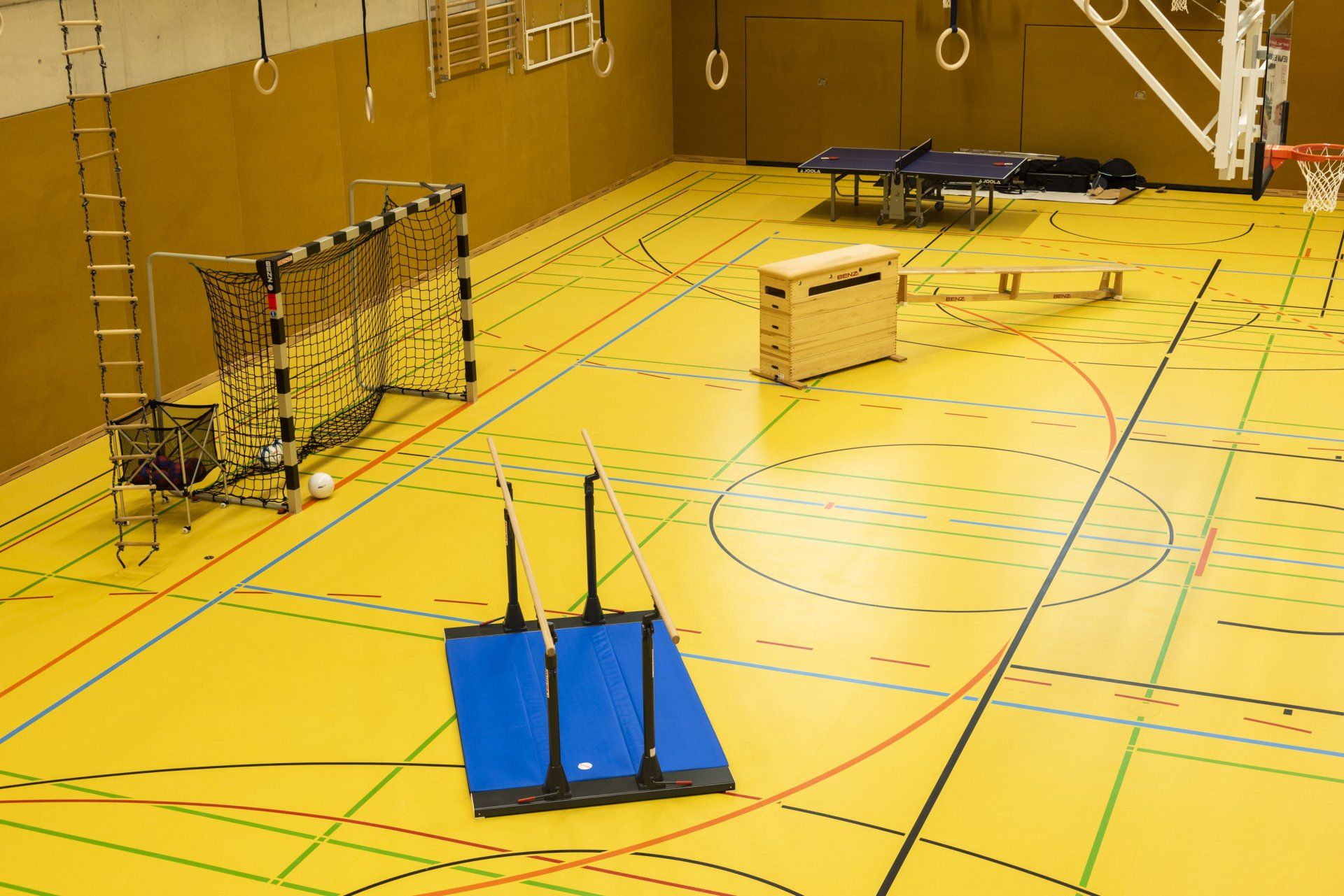 Gelber Turnhallenboden mit Sportgeräten: Fußballtor, Matten, Tische, Ringe und Leiter.