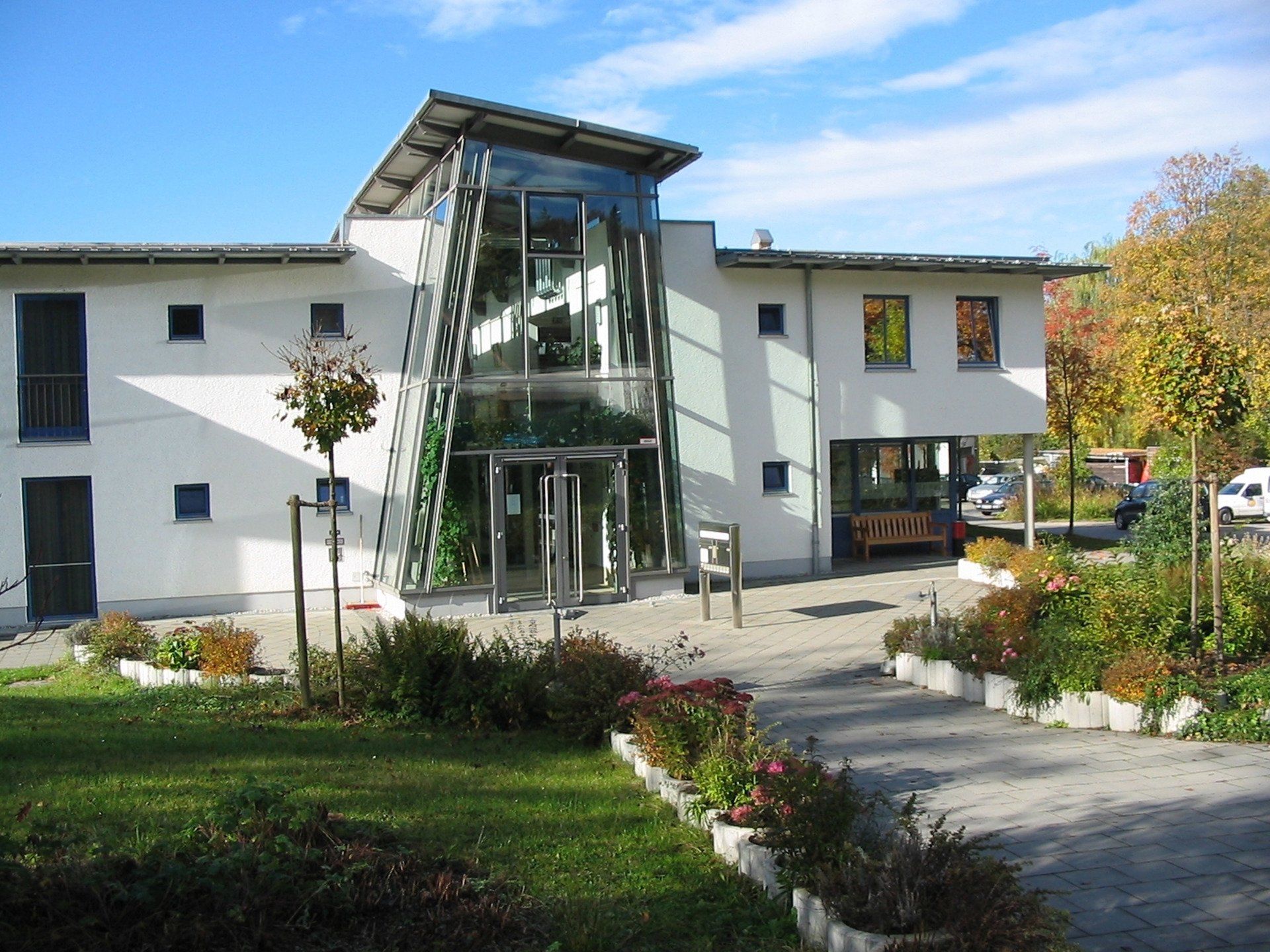 Zweistöckiges weißes Gebäude mit gläsernem Eingang, Gartenanlage und blauem Himmel.