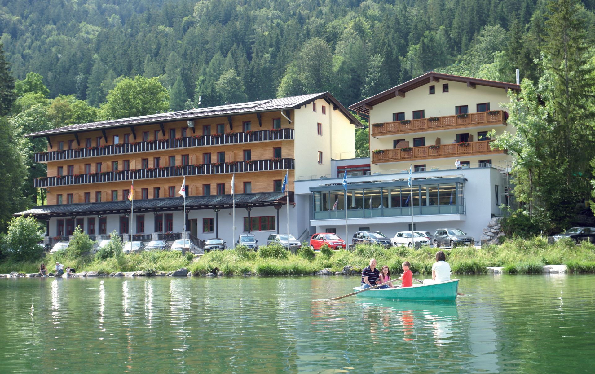 Hotel an einem See mit Ruderboot. Grünes Wasser, Bäume und bewölkter Himmel.