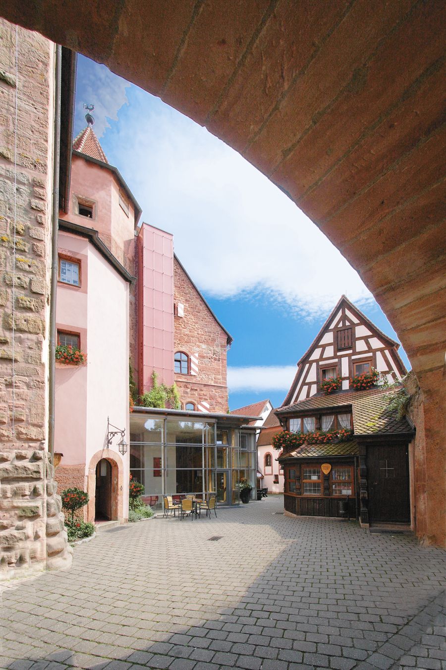Blick durch einen Torbogen auf eine Kopfsteinpflasterstraße mit rosa und braunen Gebäuden und einem Fachwerkhaus unter blauem Himmel.